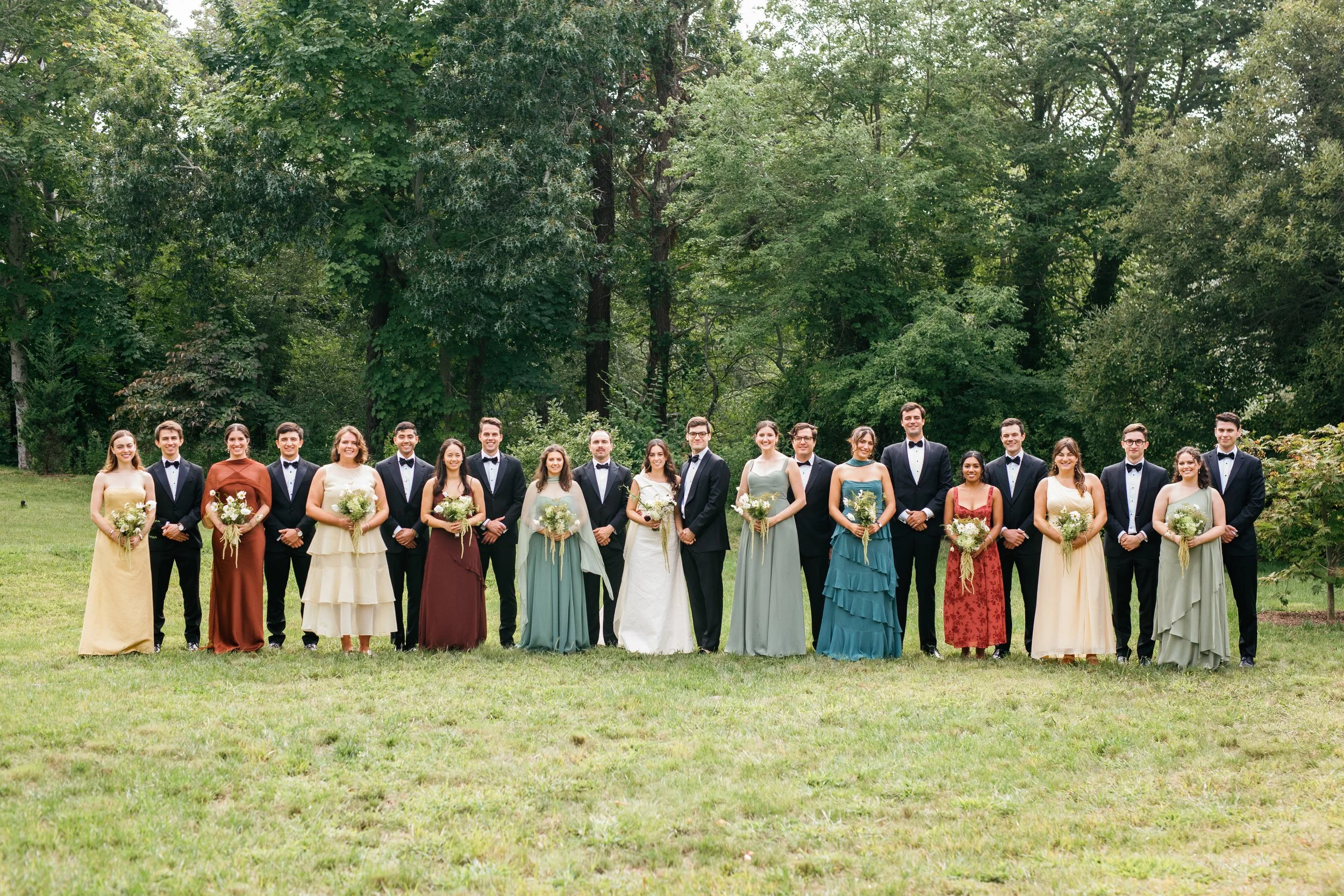 A large group of people, dressed in formal attire, standing outdoors on a grassy area with tall trees in the background, posing for a photo at a wedding or special event.