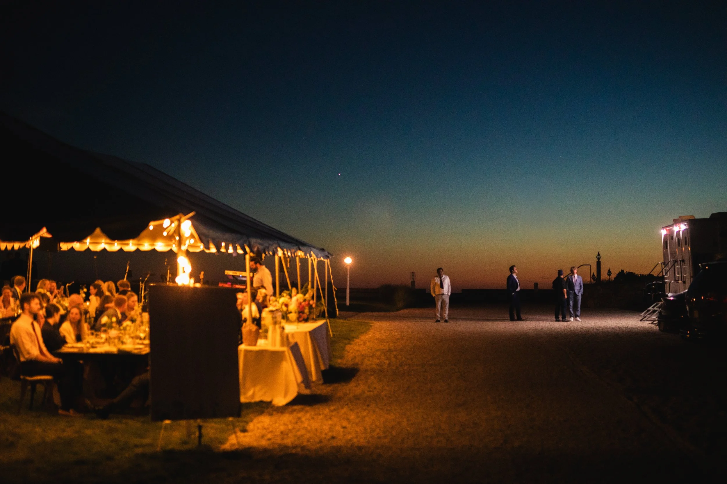 It is night time and guest enjoy wedding festivities under a tent. Some other guest wait in line for the trailer.