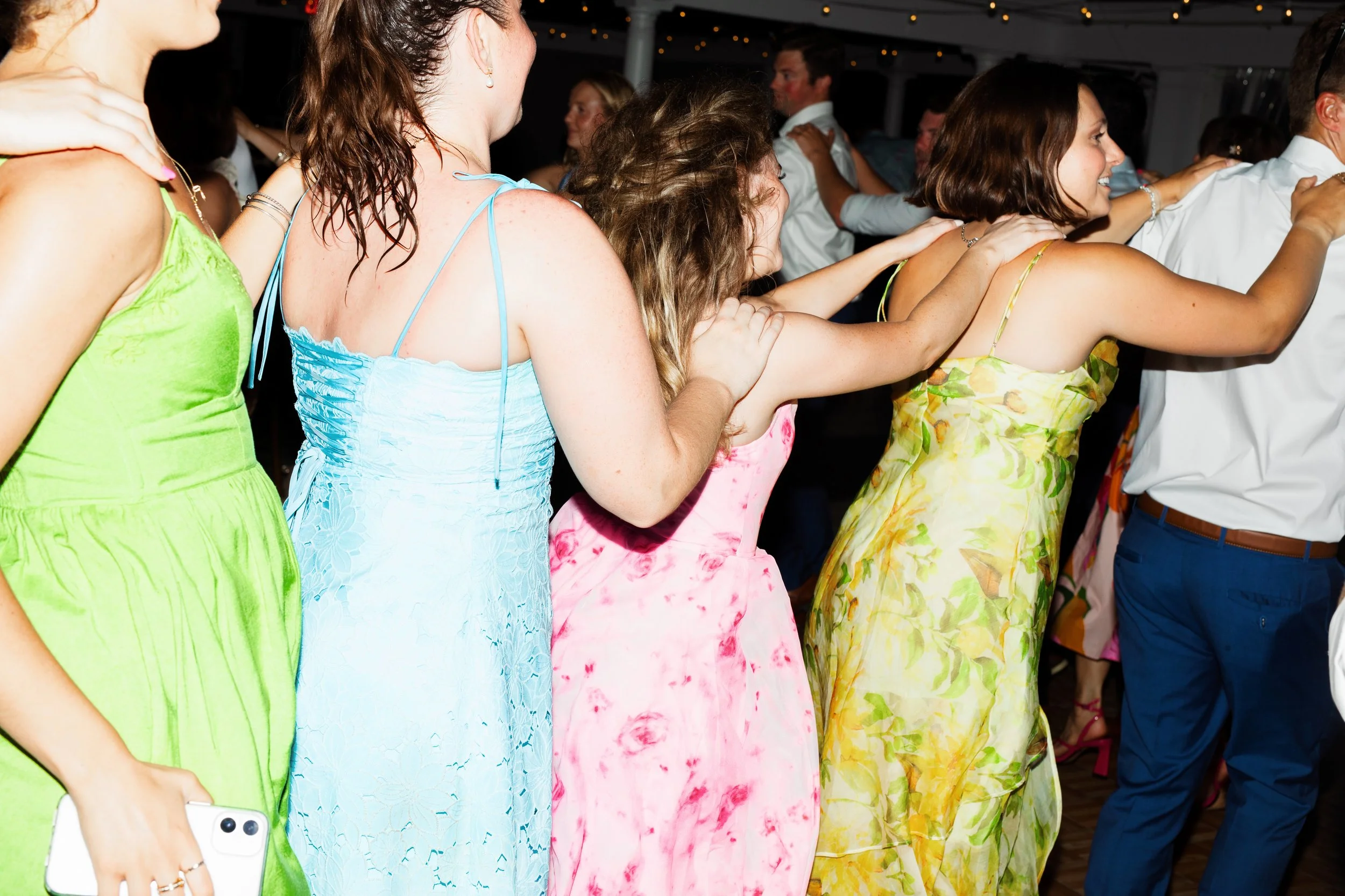 People dancing in a line at a party or celebration, wearing colorful dresses and formal attire.
