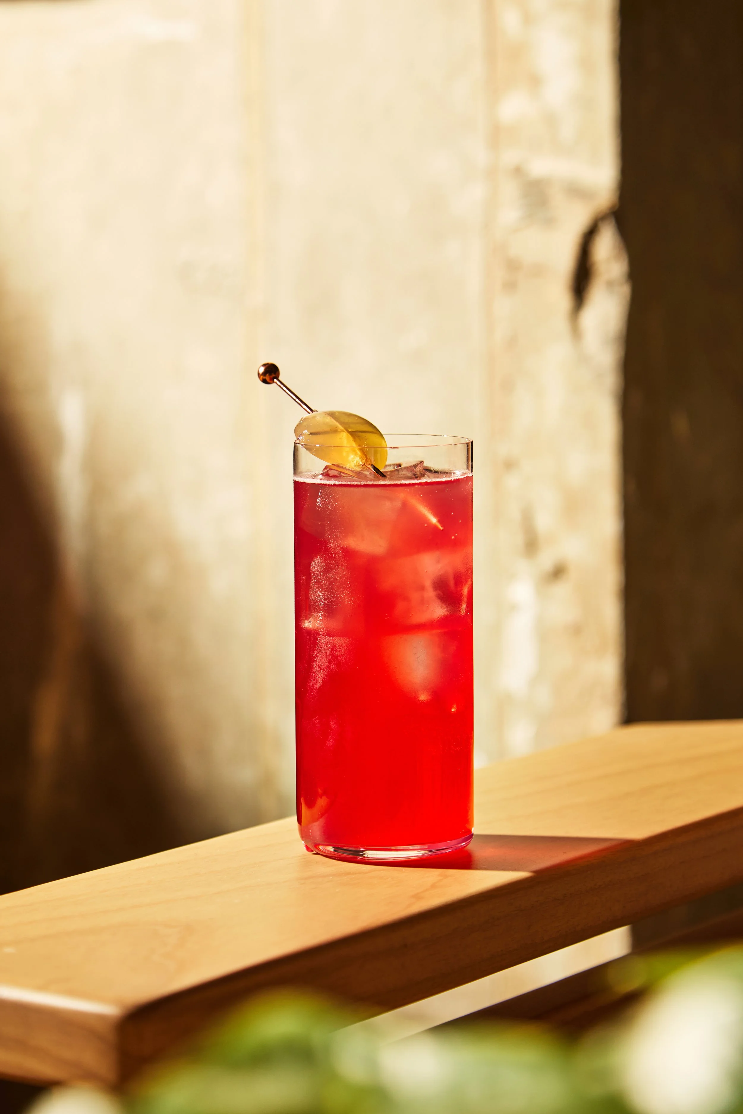 A tall glass of red cocktail with ice, garnished with a lemon slice and a cocktail pick, sitting on a wooden surface.