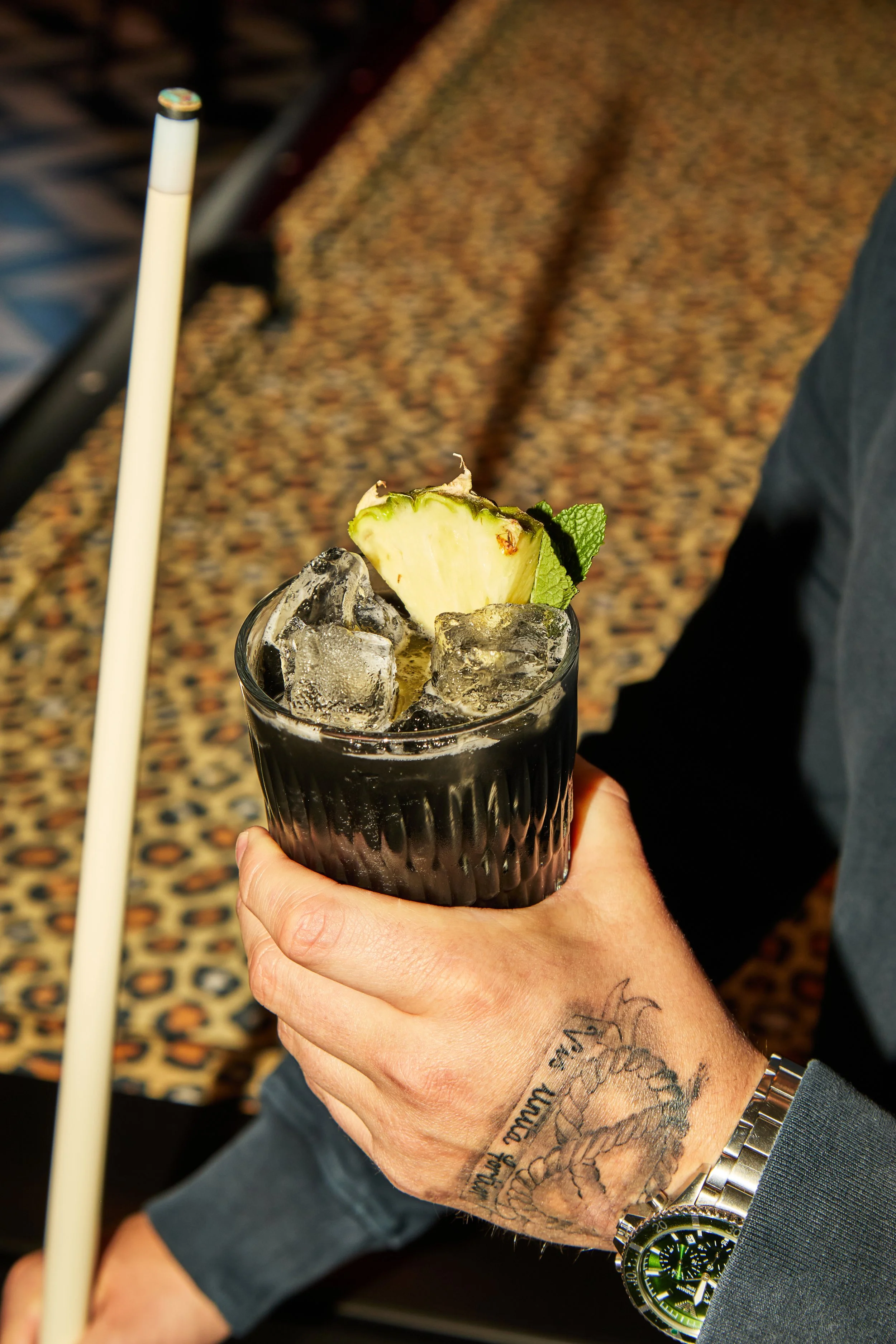 A person's hand holding a glass of dark soda with ice, garnished with a pineapple slice and mint leaf. The person has tattoos on their wrist and is wearing a silver watch. There is a white straw in the drink. The background shows a patterned carpet a