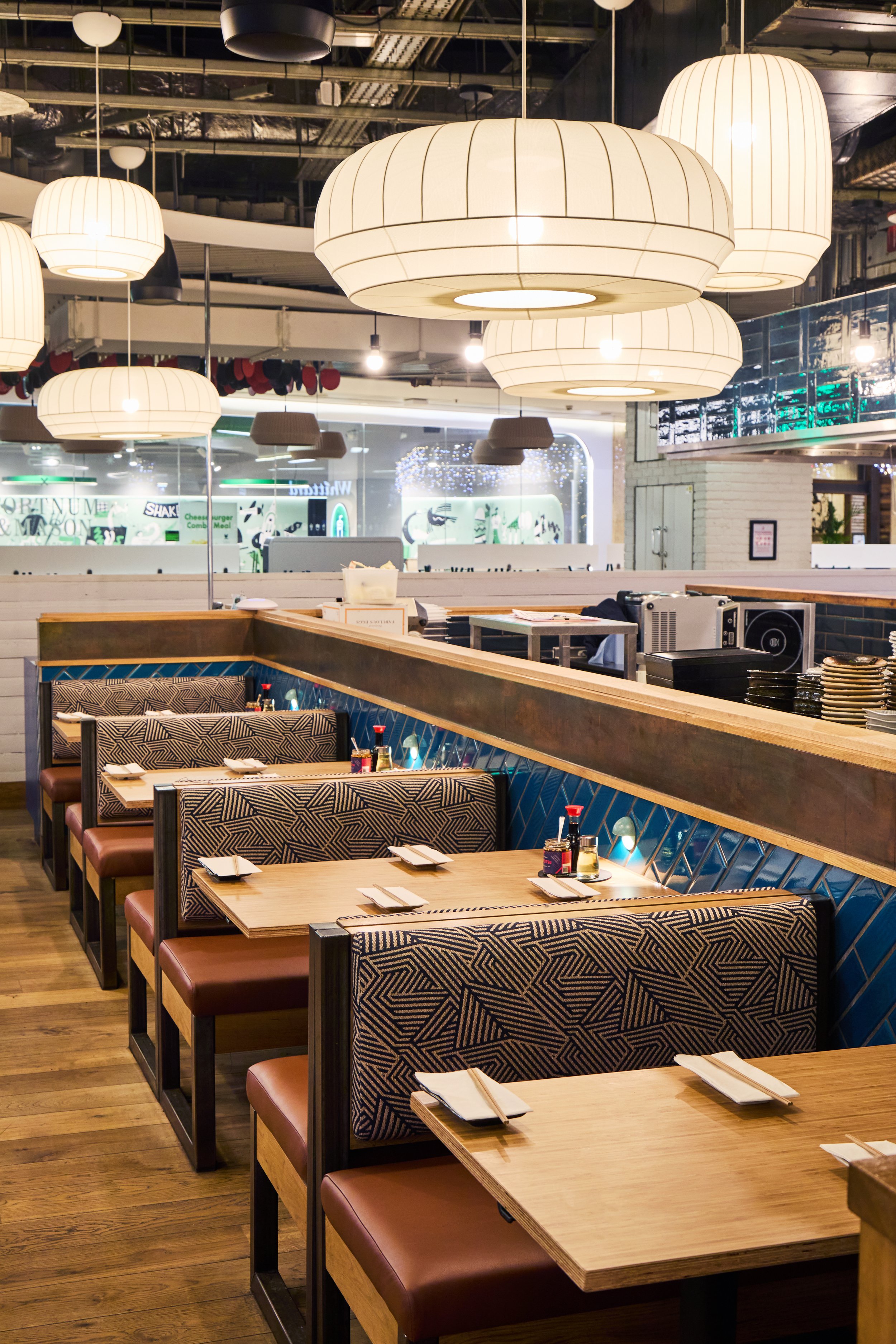 Empty sushi restaurant with tables, patterned booths, chopsticks, soy sauce, and large hanging white lamps.