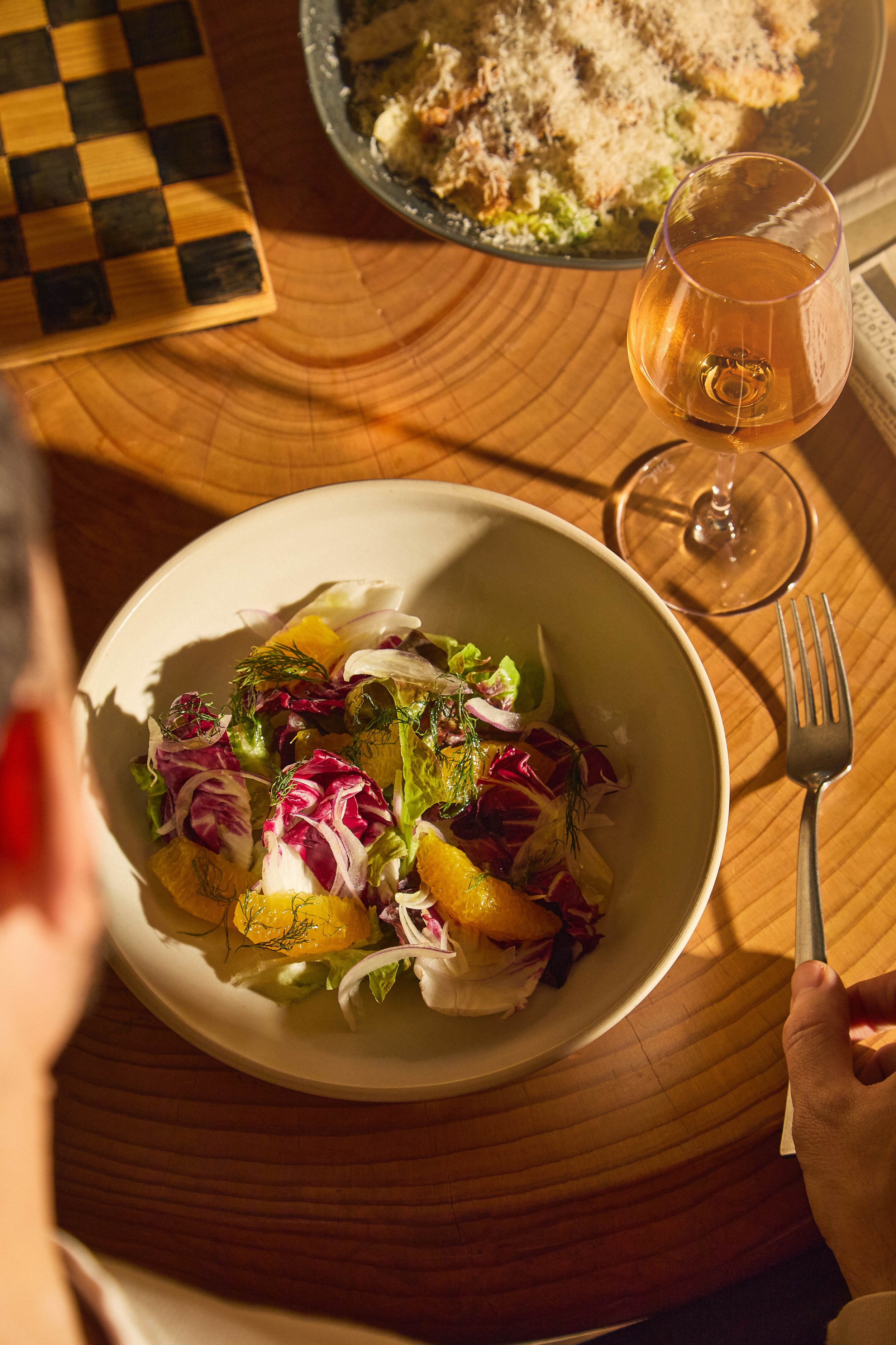 A wooden table with a checkered pattern, a bowl of mixed salad with greens and orange slices, a glass of rosé wine, and a plate with fried food and a fork.