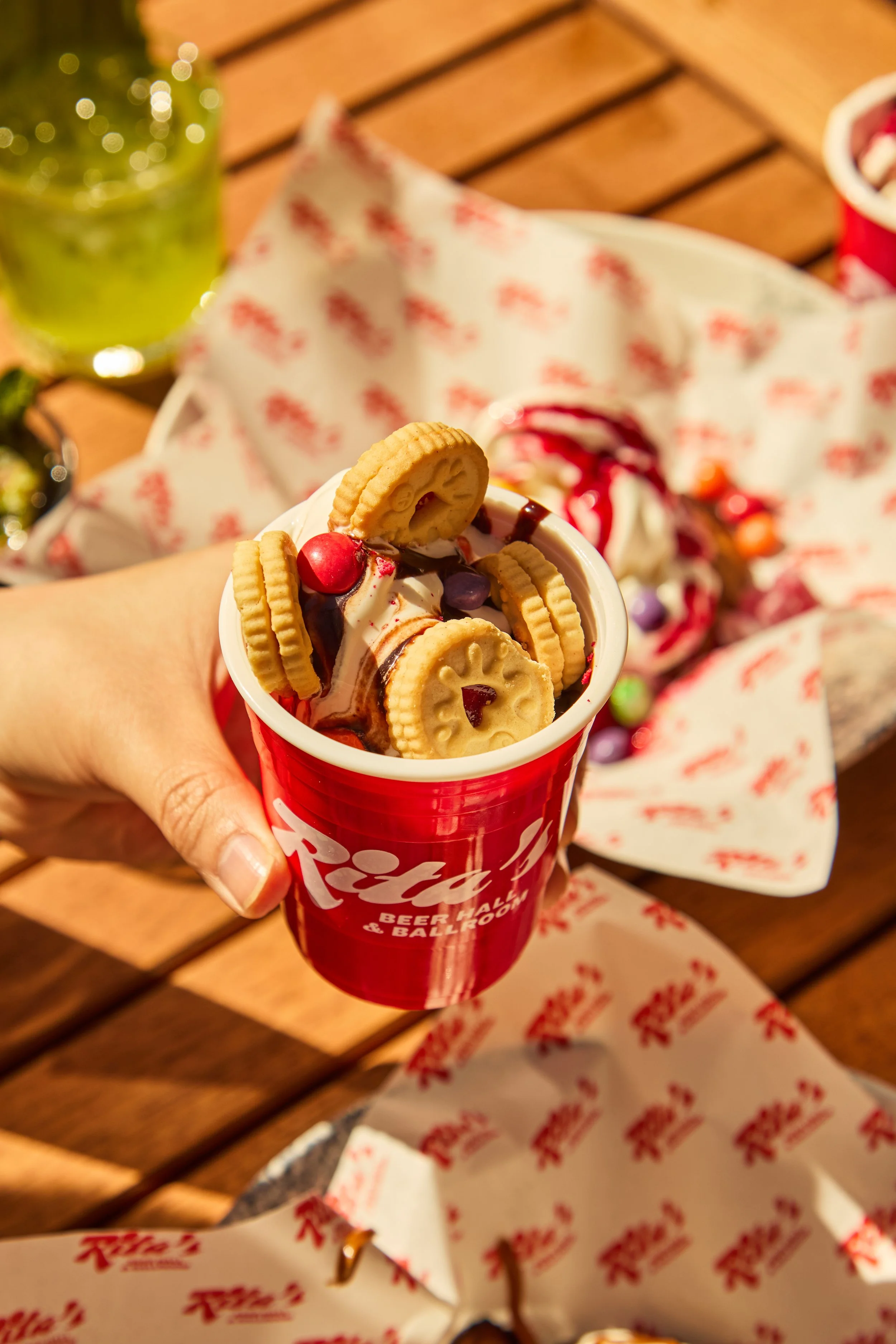 A person holding a red cup filled with ice cream, Oreo cookies, and colorful candies, with a Dr. Pepper logo on the cup. There are Oreo cookies, a chocolate drizzle, and candies on top, and a green drink and red popcorn bucket are visible in the back