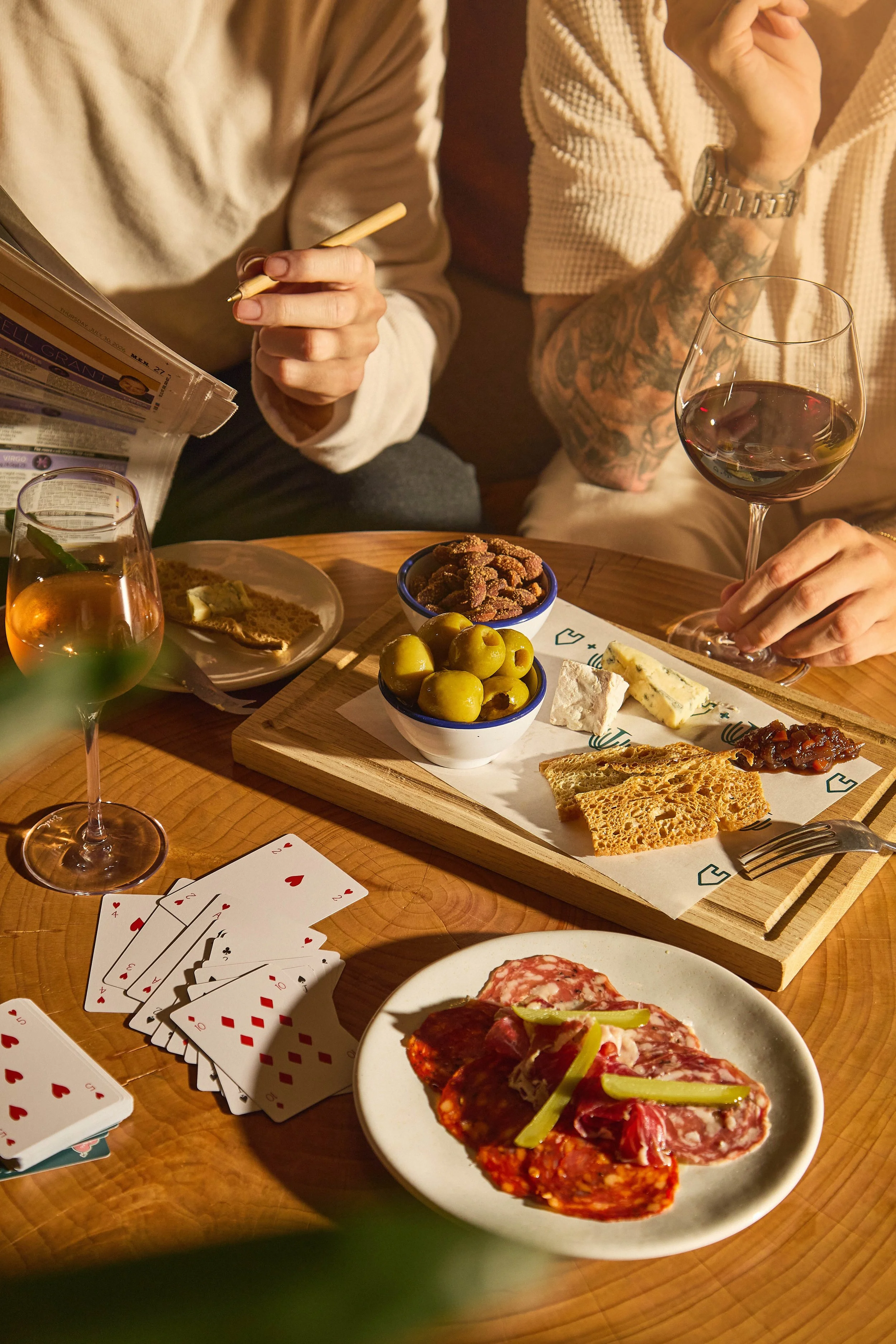 A table set for a gathering with various foods including sliced cheese, olives, cured meats, and slices of bread or crackers. There are also glasses of red wine and a plate of pizza. Playing cards are spread out on the table, indicating a social game