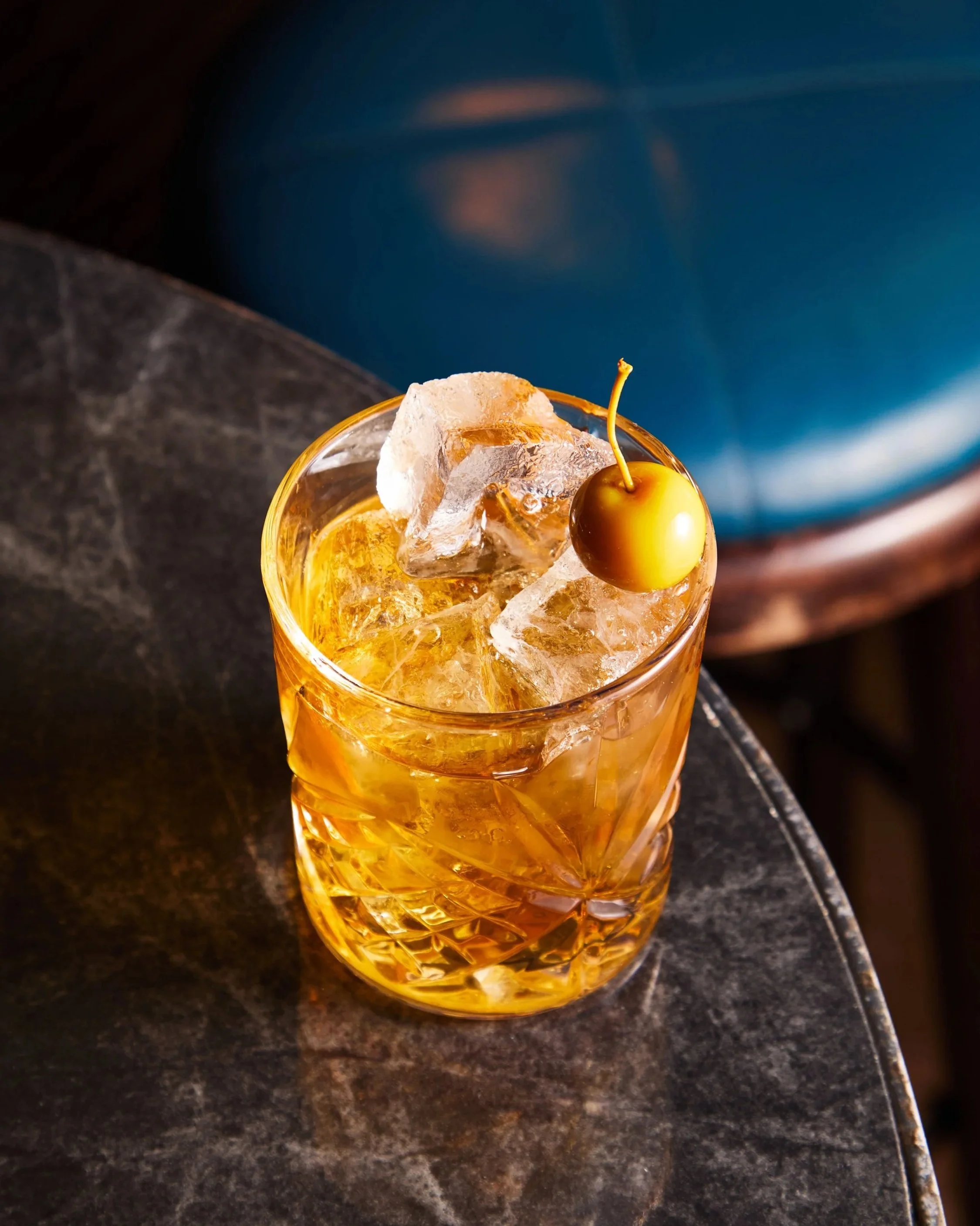 A glass of whiskey with ice and a cherry on top placed on a dark textured table.