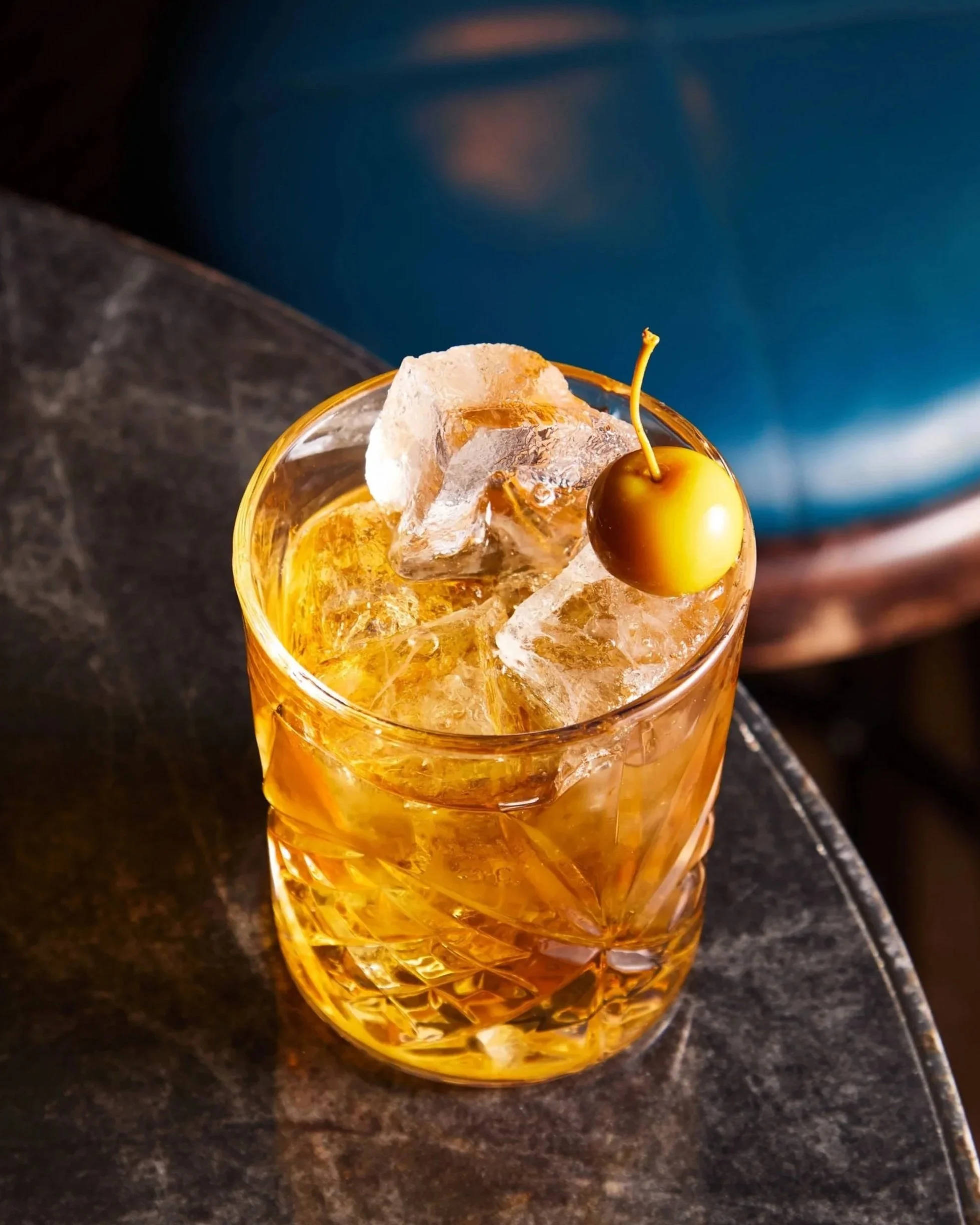 A glass of whiskey with ice and a cherry on top placed on a dark textured table.