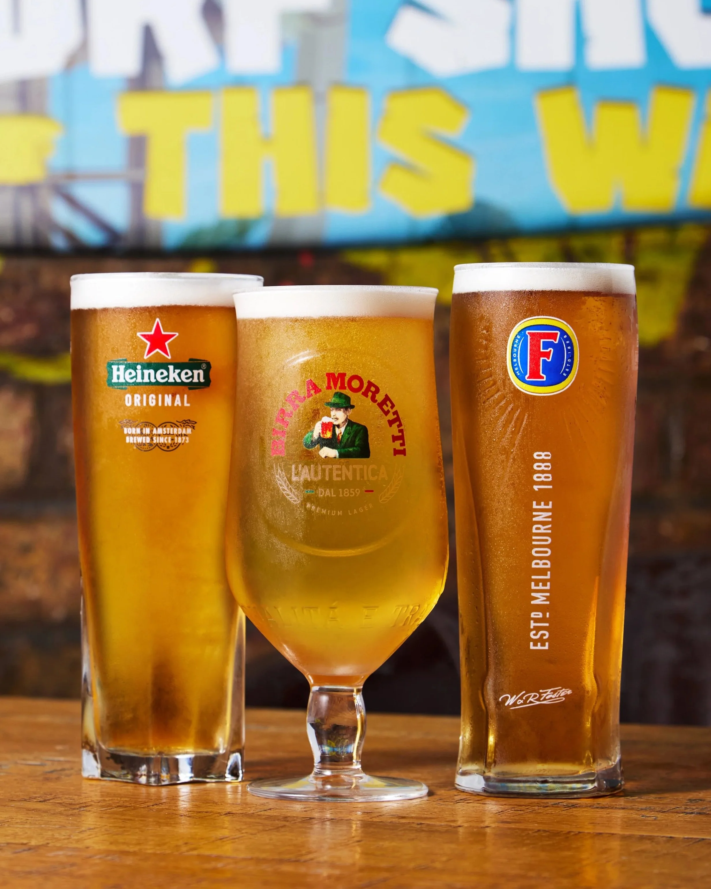 Three glasses of beer on a wooden surface with a colorful sign in the background. The left glass is a tall Heineken beer, the middle glass is a branded beer with a red logo, and the right glass is a tall Fosters beer.