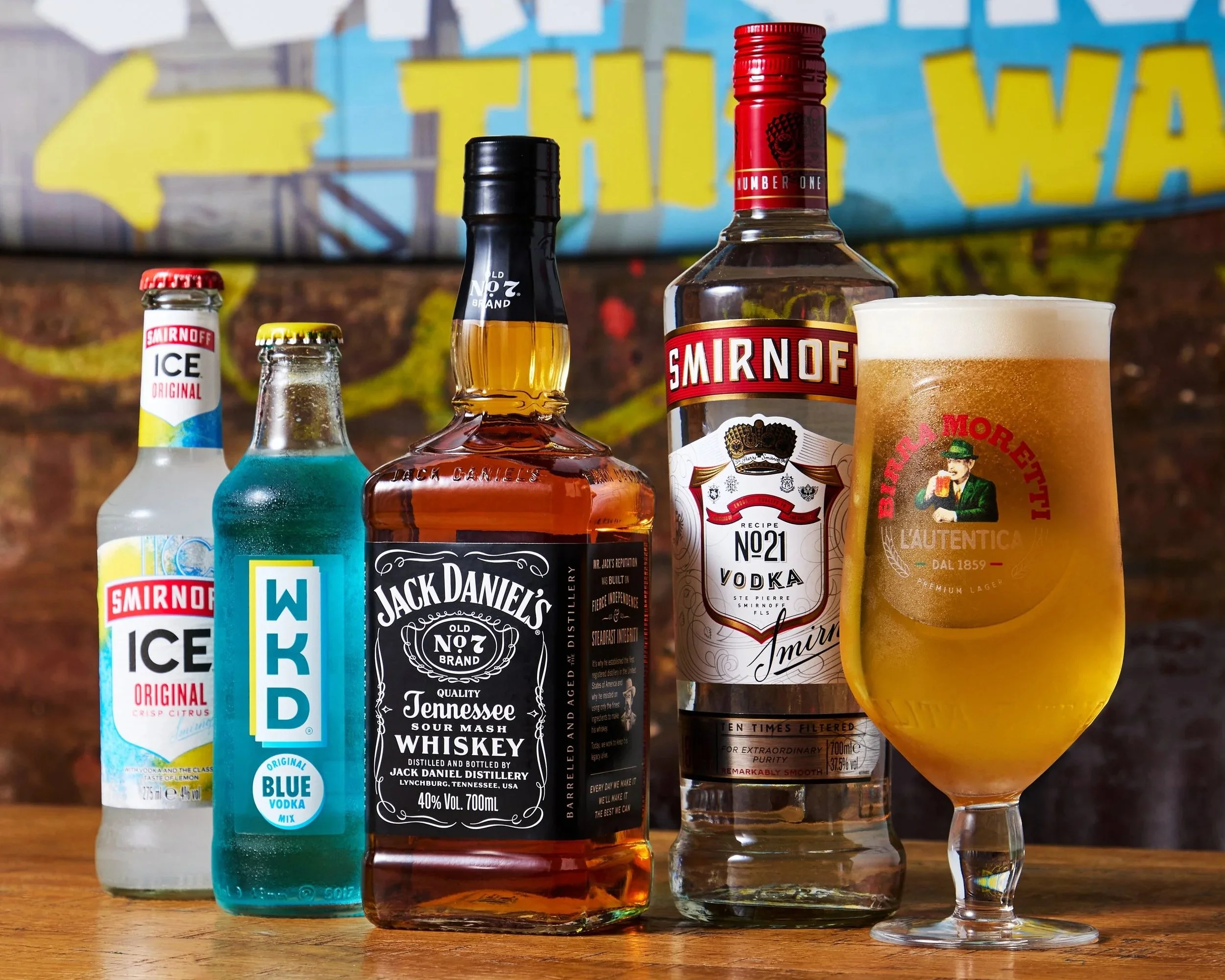 Various alcoholic beverages including Smirnoff Ice, WKD Blue Vodka Mix, Jack Daniel's Tennessee Whiskey, Smirnoff No. 21 Vodka, and a glass of beer, displayed on a wooden surface with a colorful background.
