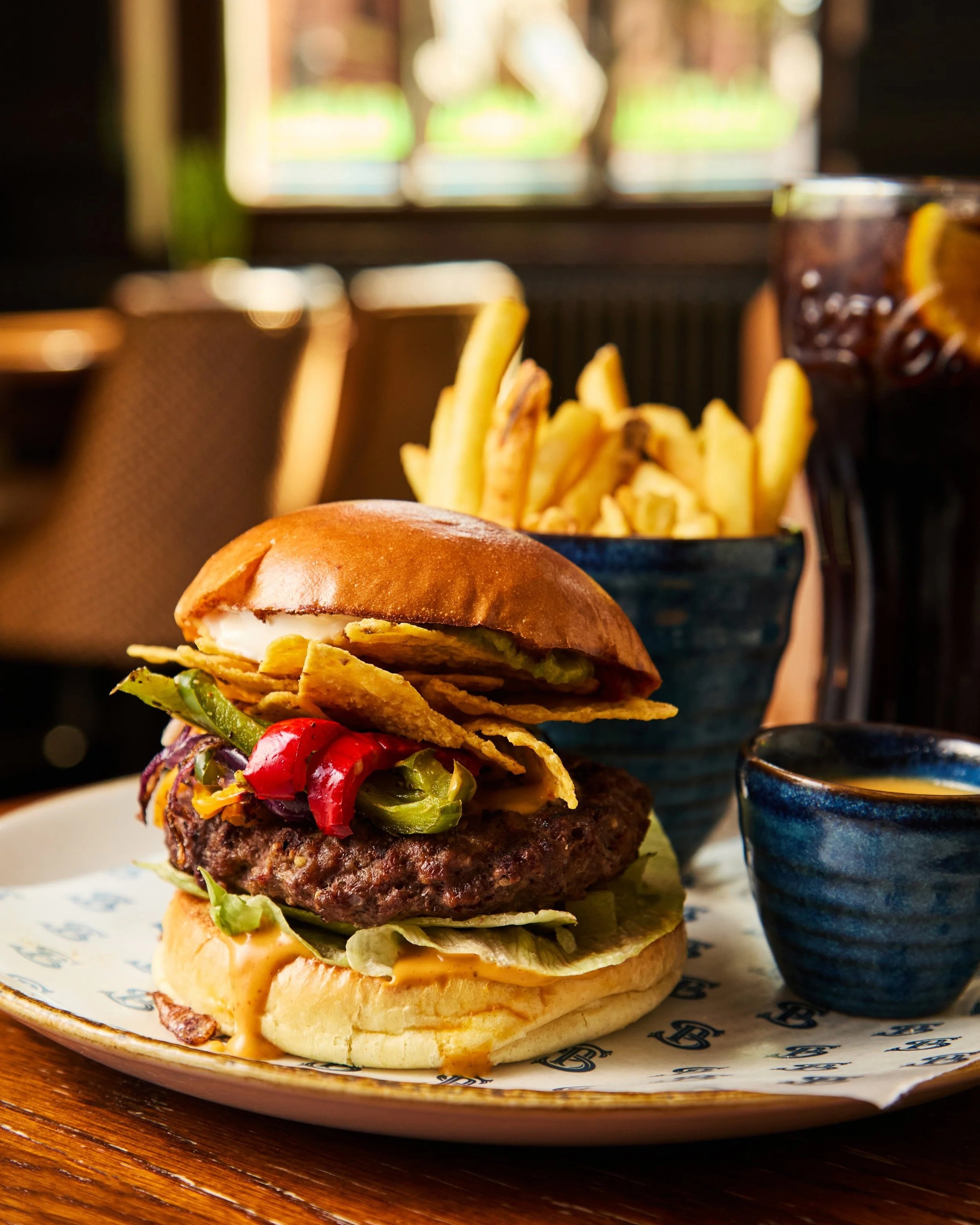 Close-up of a gourmet cheeseburger with lettuce, tomato, pickles, and grilled onion in a bun, served on a decorative plate with a side of French fries and a dark soda in a glass. The background shows a blurred window with natural light.