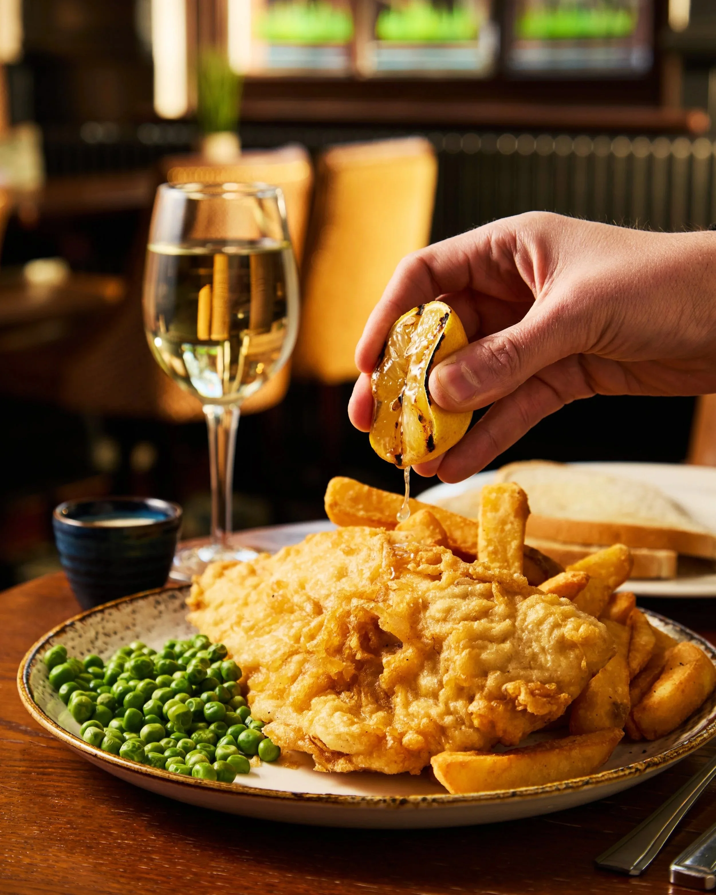 Fried fish fillets with green peas and French fries on a decorative plate, with a glass of sparkling water and a small black dipping sauce, set on a wooden table in a restaurant.