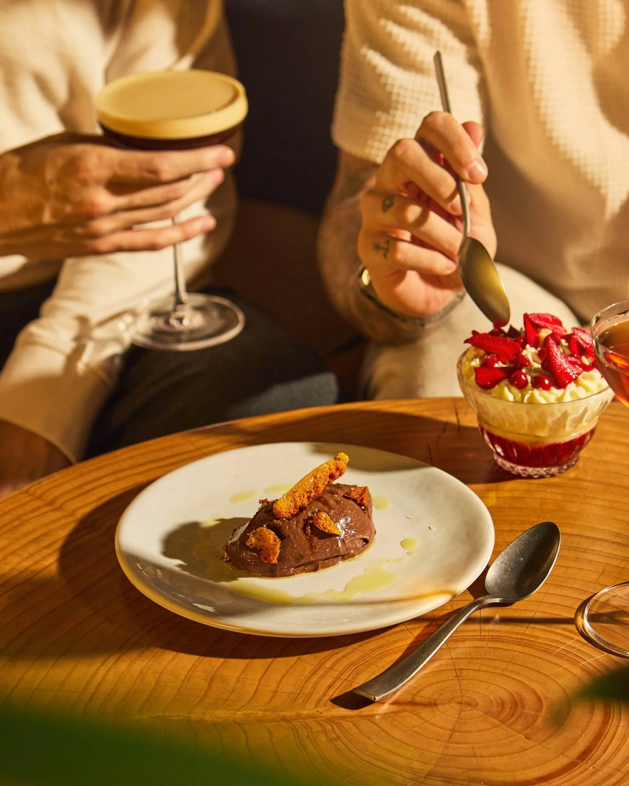 A dessert with a scoop of chocolate ice cream garnished with cookie crumbles on a white plate, placed on a wooden table. In the background, a woman is holding a spoon and a bowl of strawberry shortcake with strawberries and whipped cream, and a man i