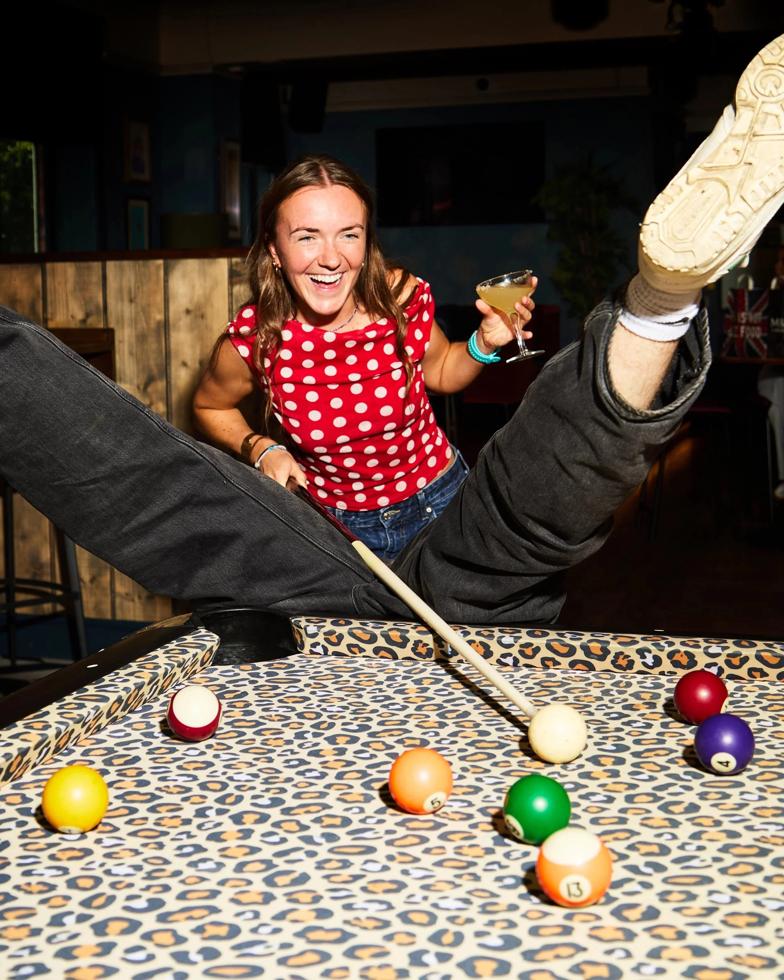 A young woman smiling and laughing while holding a cocktail, sitting next to a pool table with colorful billiard balls.