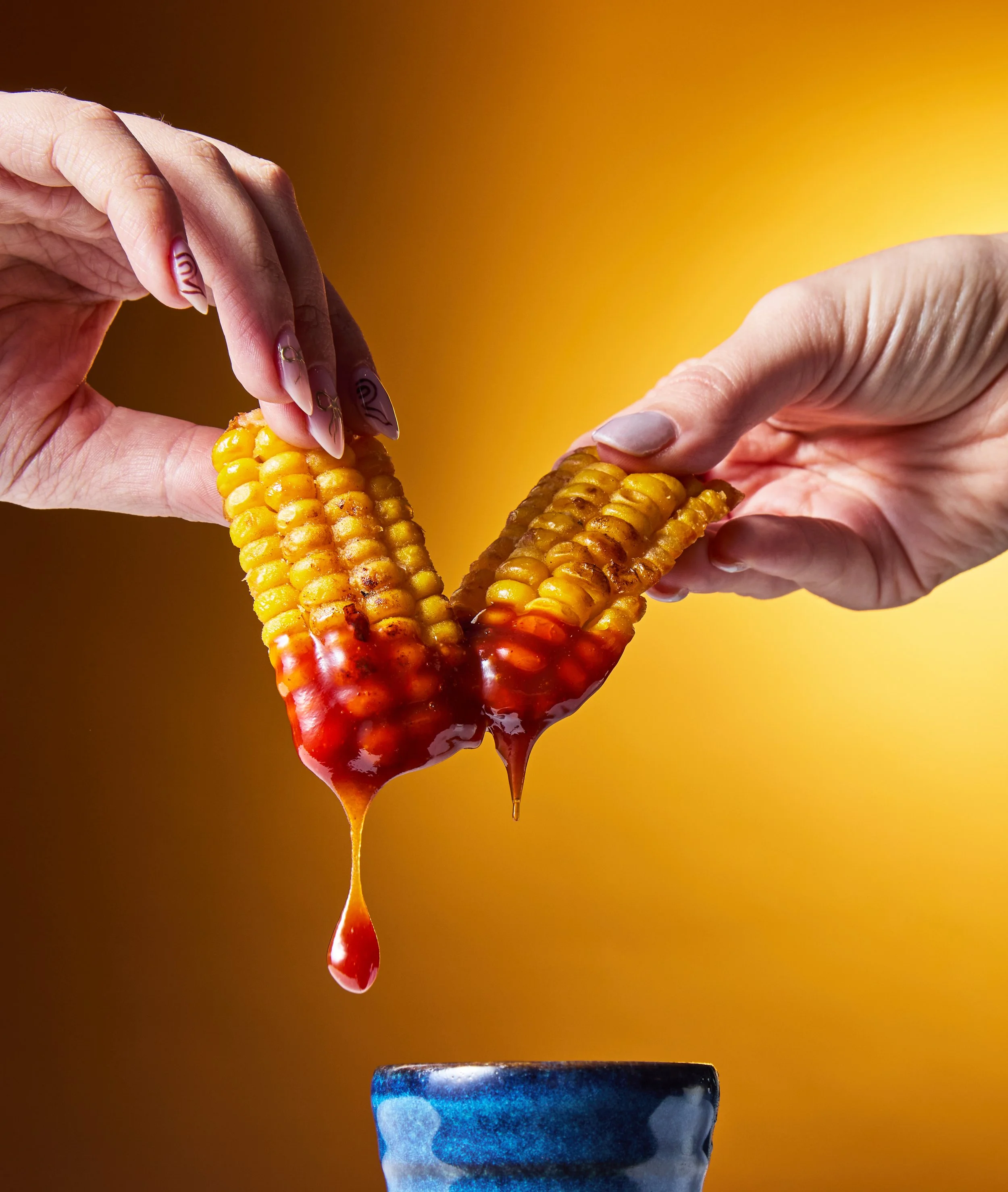 Two hands holding grilled corn on the cob dipped in ketchup, dripping, with a blue ceramic cup below.