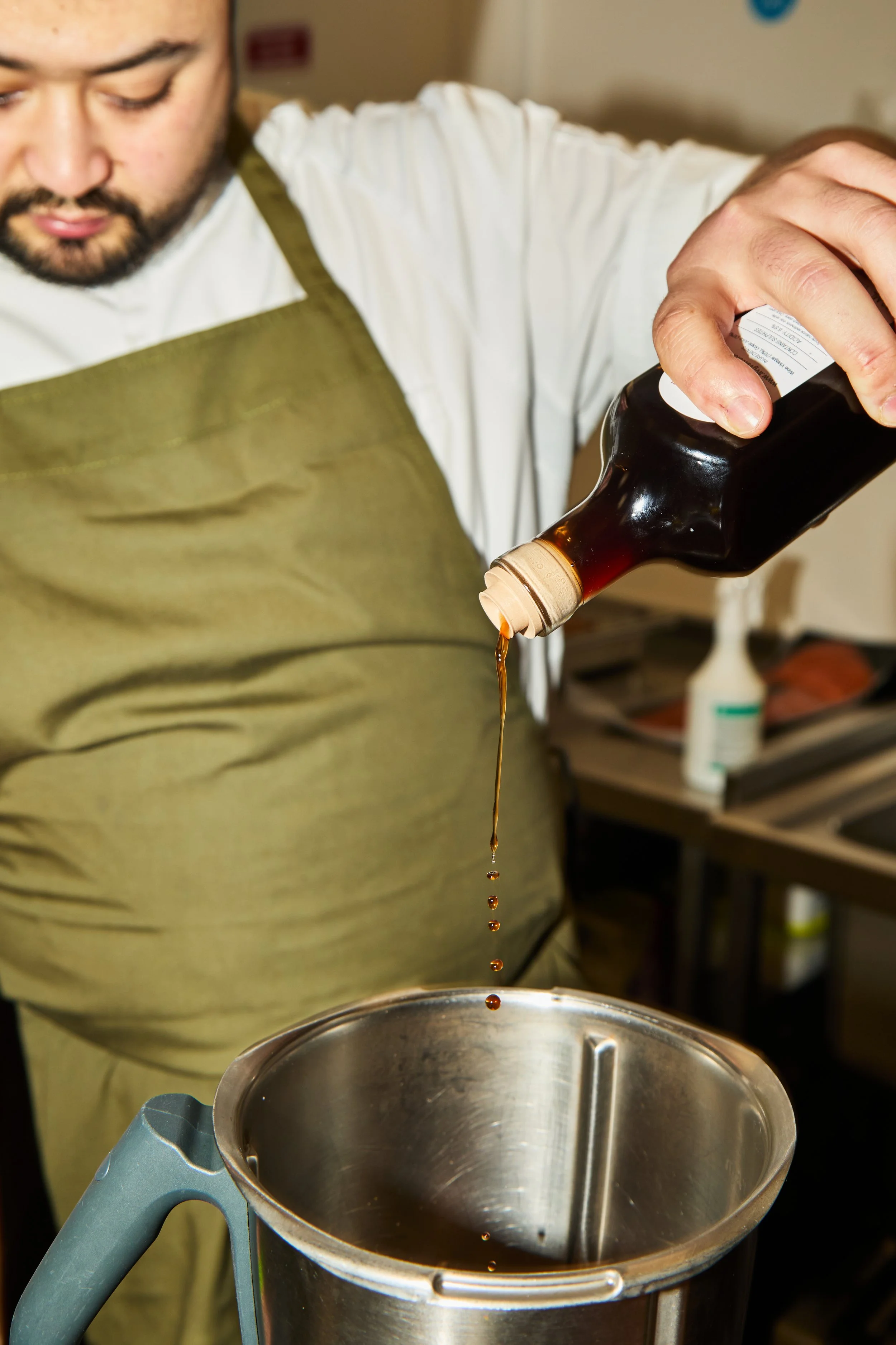 A person pouring soy sauce from a bottle into a stainless steel container.