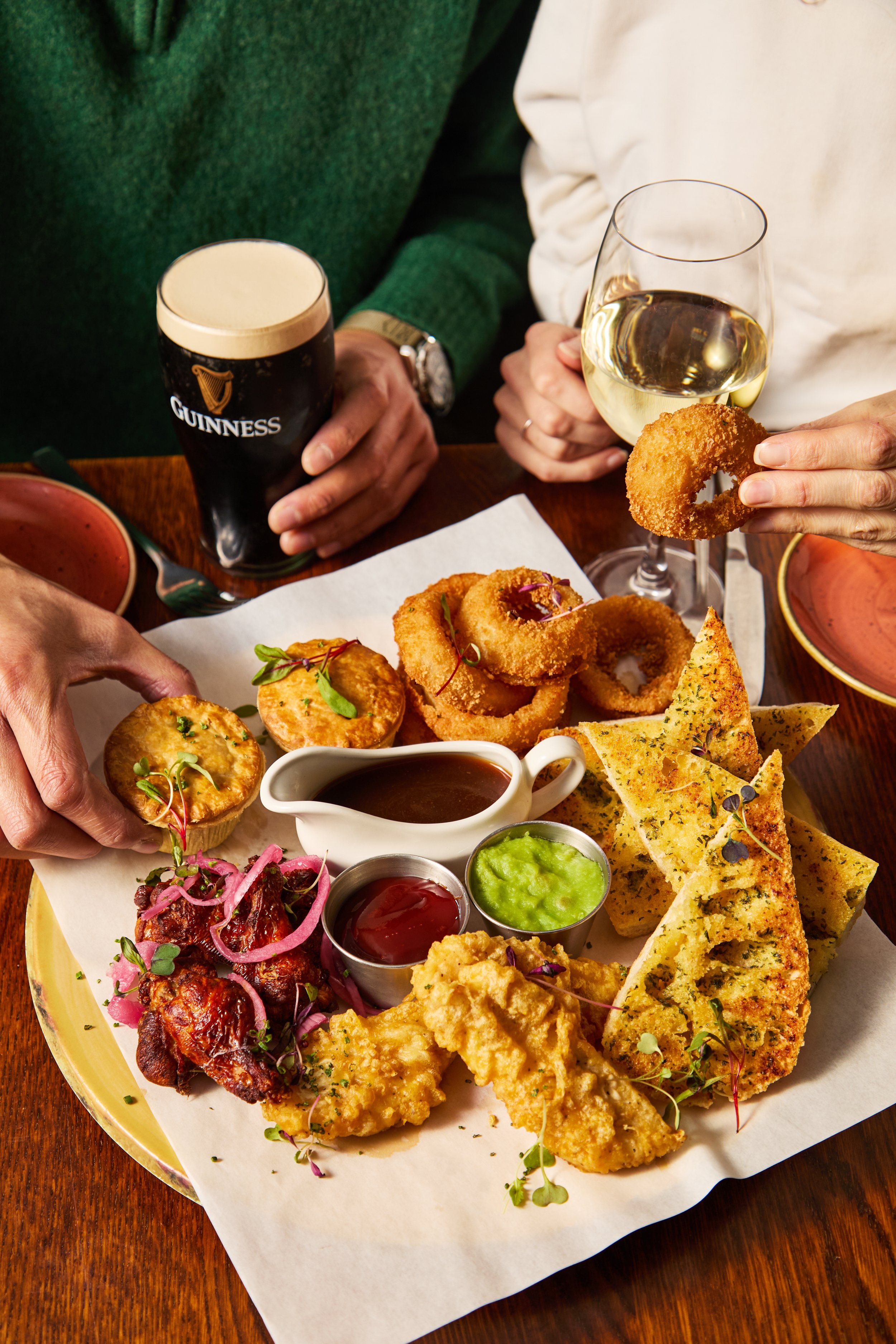 People enjoying a platter of assorted fried foods, onion rings, chicken wings, and breaded appetizers with dipping sauces, while holding drinks of Guinness beer and white wine.