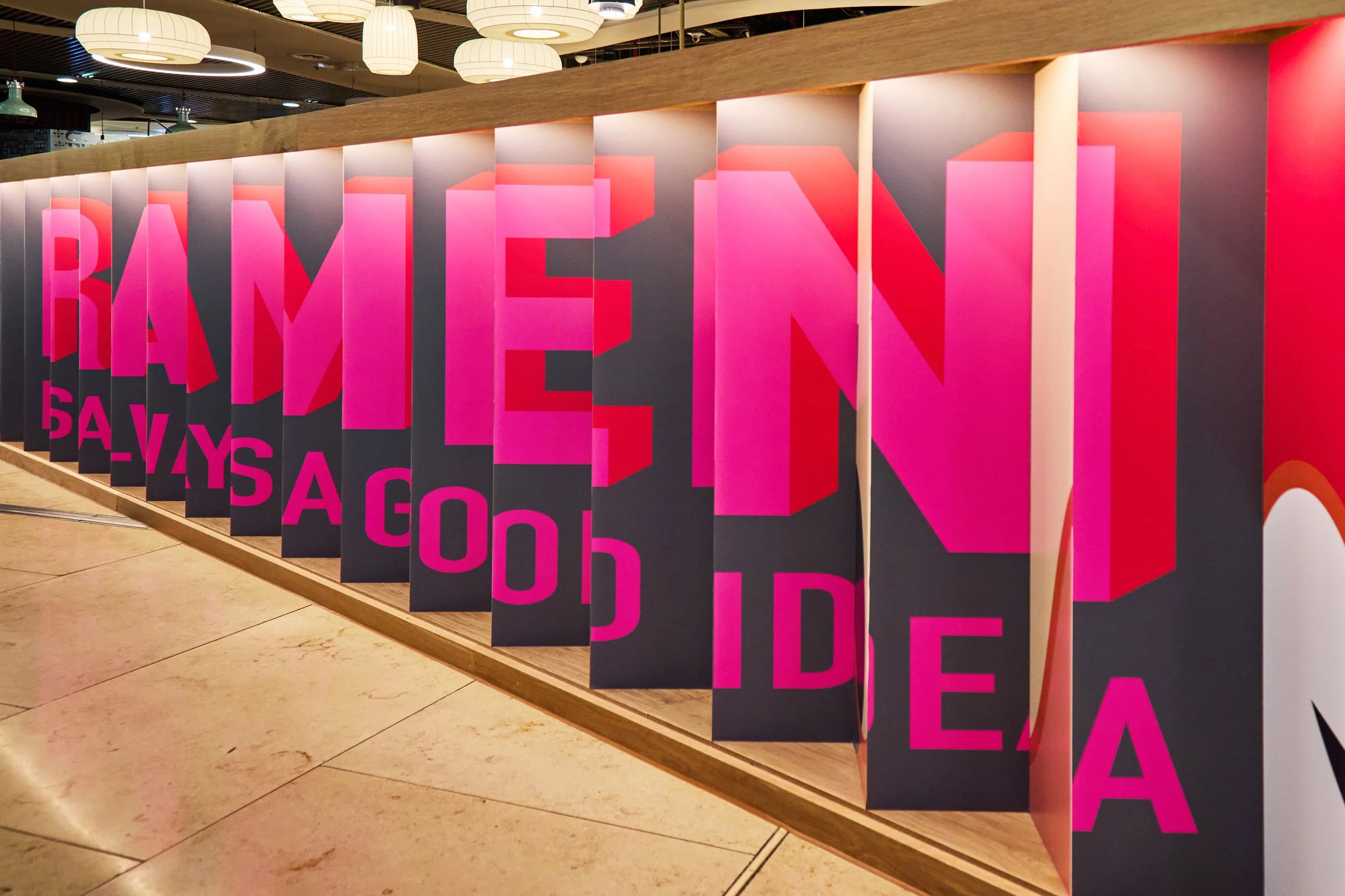 Colorful display of large, pink and red 3D letters reading 'RAMEN' and the smaller words 'ALWAYS A GOOD IDEA' on black vertical panels in a modern setting.