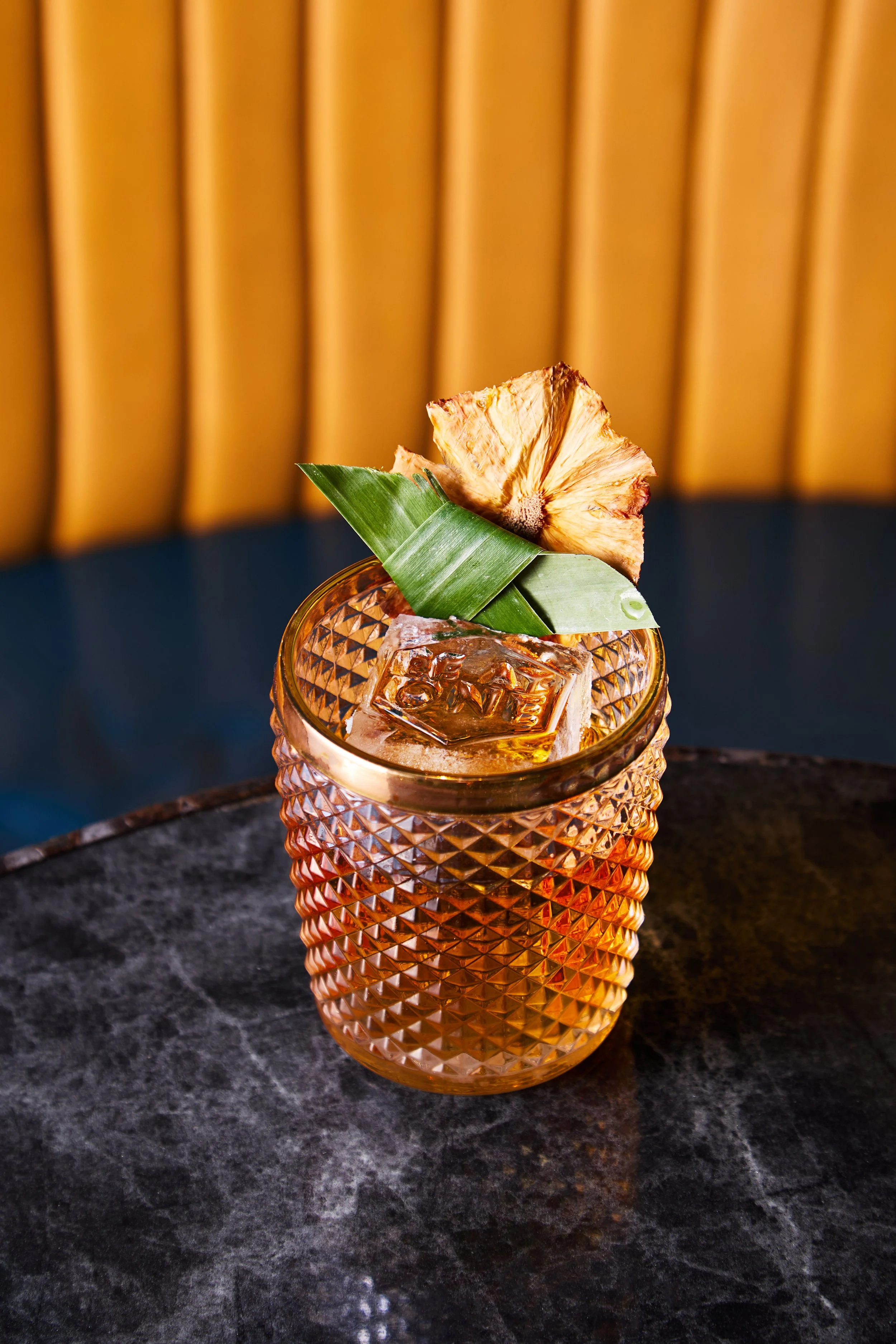 A textured glass filled with a dark cocktail, garnished with a pineapple slice, a green leaf, and a large ice cube, on a dark marble surface with a yellow-tan padded background.