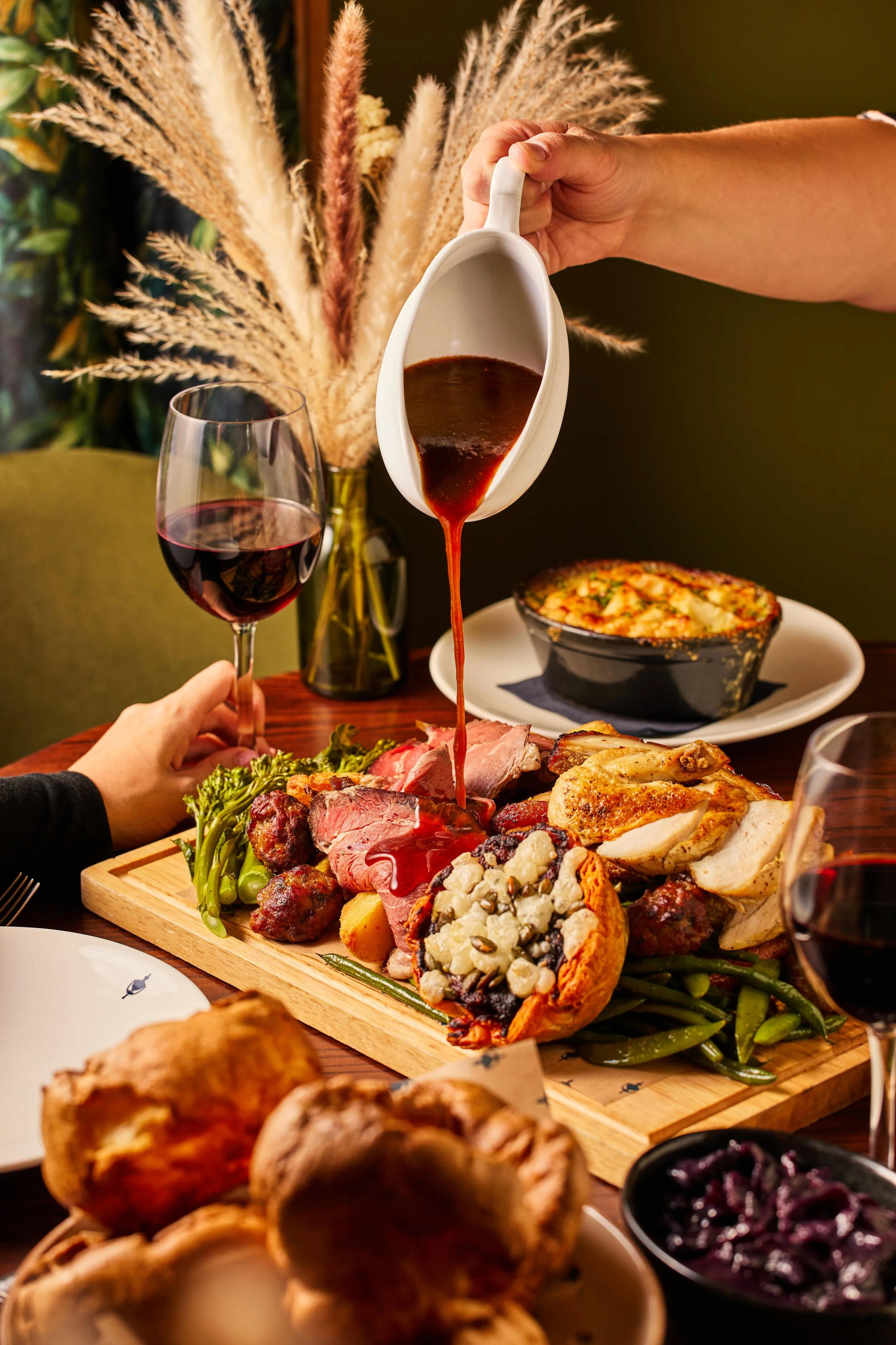 A serving of red wine in a glass, a tray of assorted meats and vegetables, a bowl of dark berries, and a black bowl of blueberries on a wooden table with a vase of dried grasses in the background.
