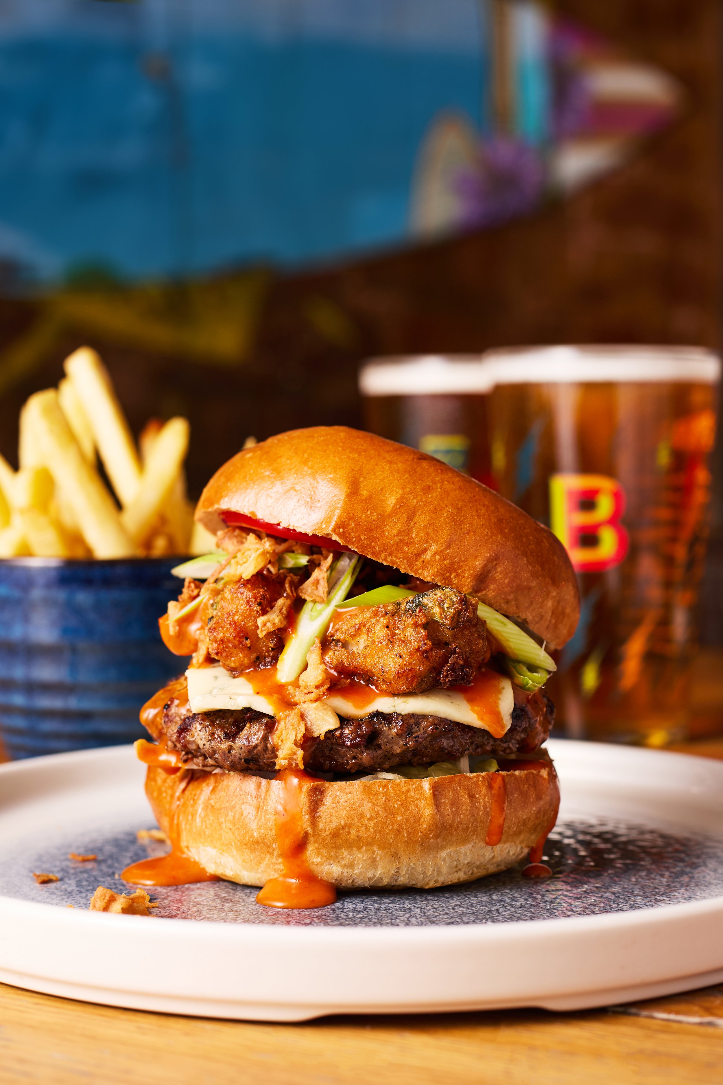 A close-up of a gourmet burger with a beef patty, fried chicken, cheese, lettuce, and sauce in a toasted bun, served on a white plate with a side of French fries and two glasses of beer in the background.