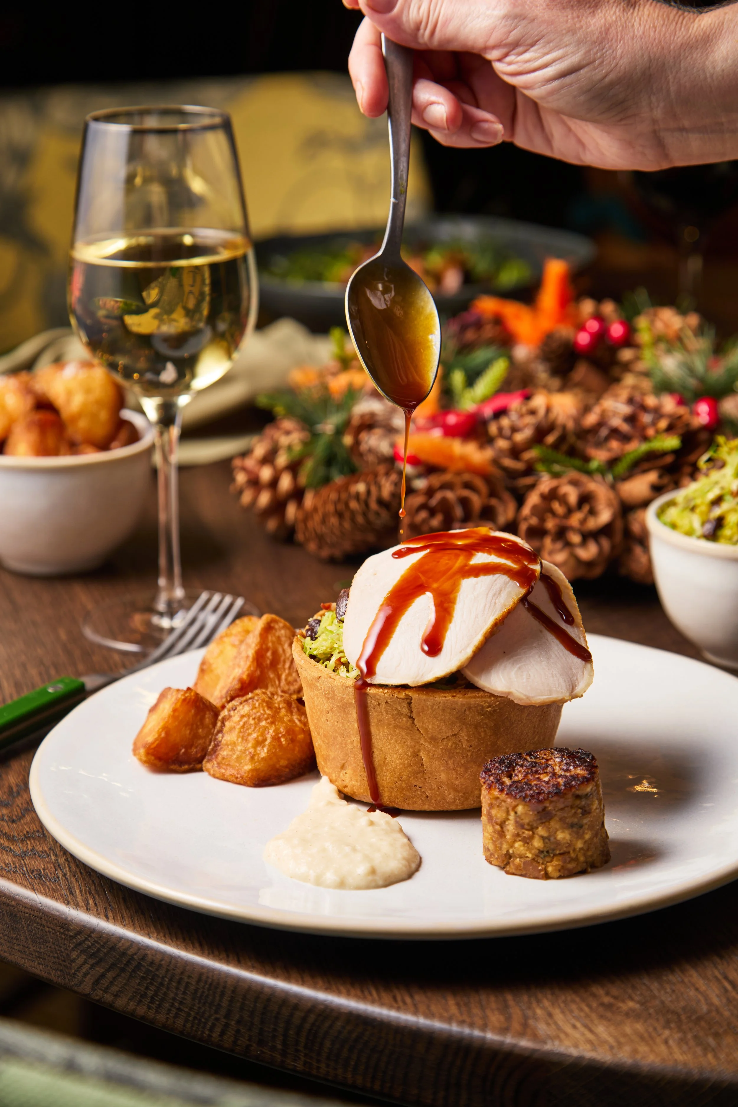 A plate with a pumpkin-shaped bread bowl filled with salad or a dish topped with sliced turkey, sauce being drizzled from a spoon, alongside roasted potatoes, a dollop of sauce, and a small cylindrical side item. In the background, a glass of white w