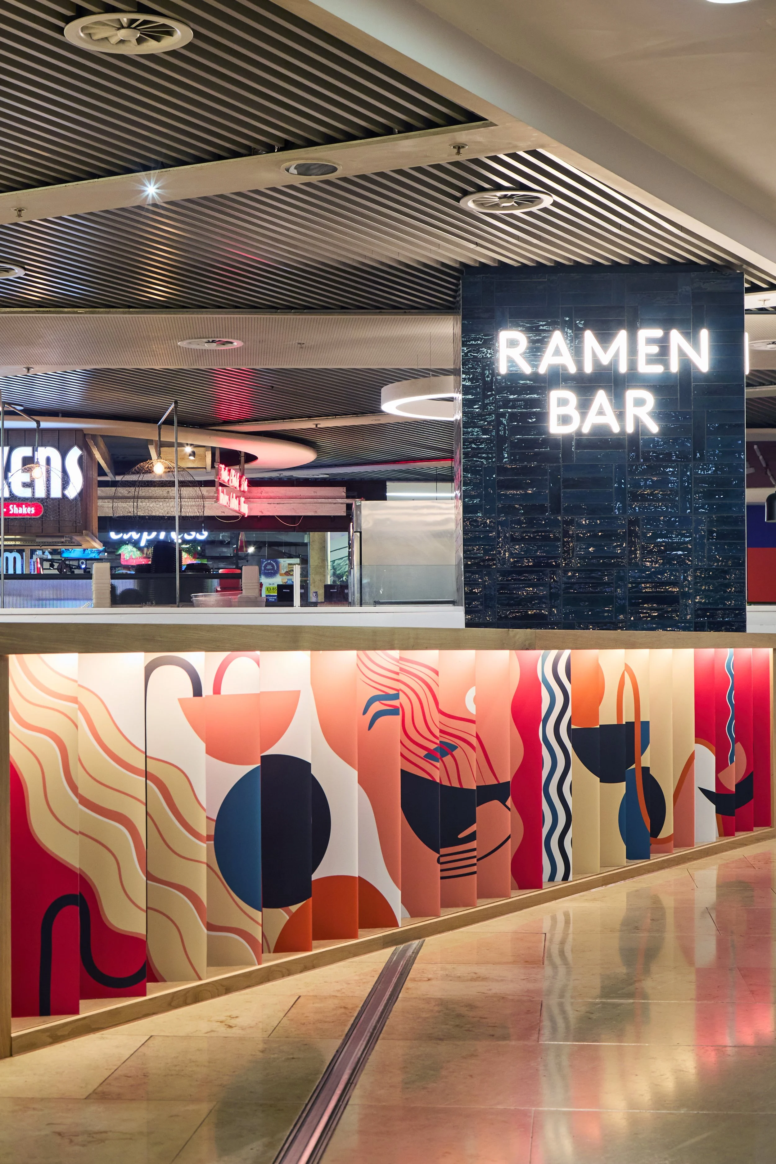 Neon sign reading 'Ramen Bar' inside a shopping mall or food court with colorful abstract art on the divider.