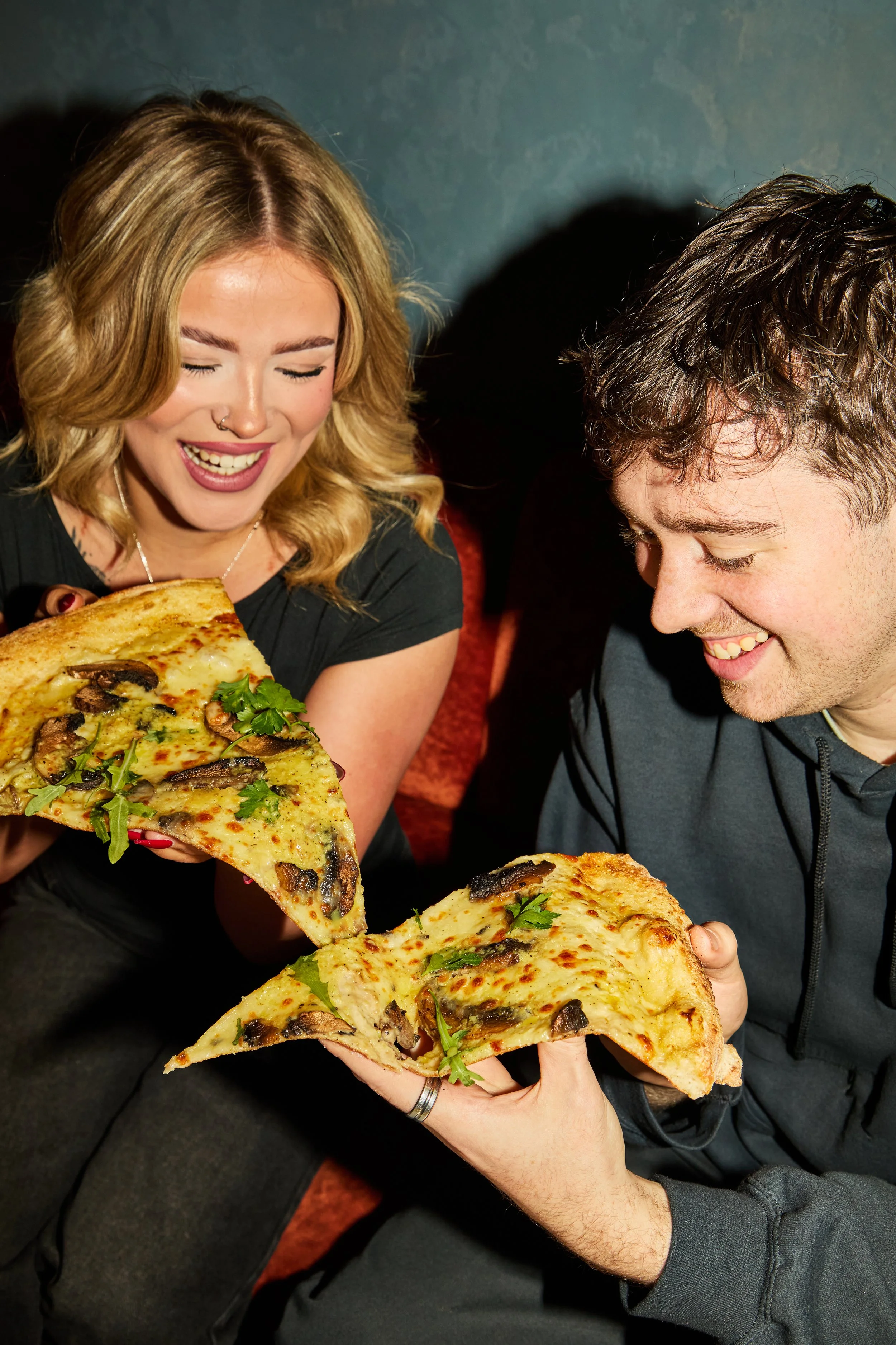 A woman and a man happily sharing pizza. The woman has blonde hair, a nose ring, and is smiling with eyes closed, holding a slice of mushroom and green garnish pizza. The man has dark hair, a dark hoodie, and is smiling, holding a slice of pizza topp