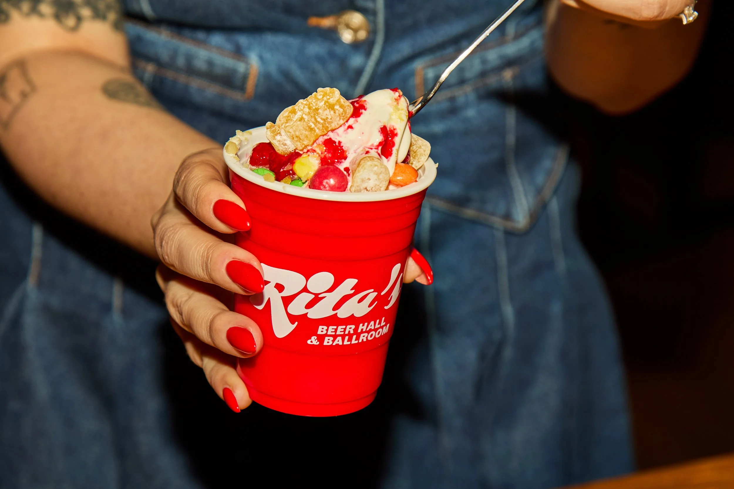 Person holding a red cup filled with ice cream topped with candies, syrup, and a cookie, in front of a denim shirt.