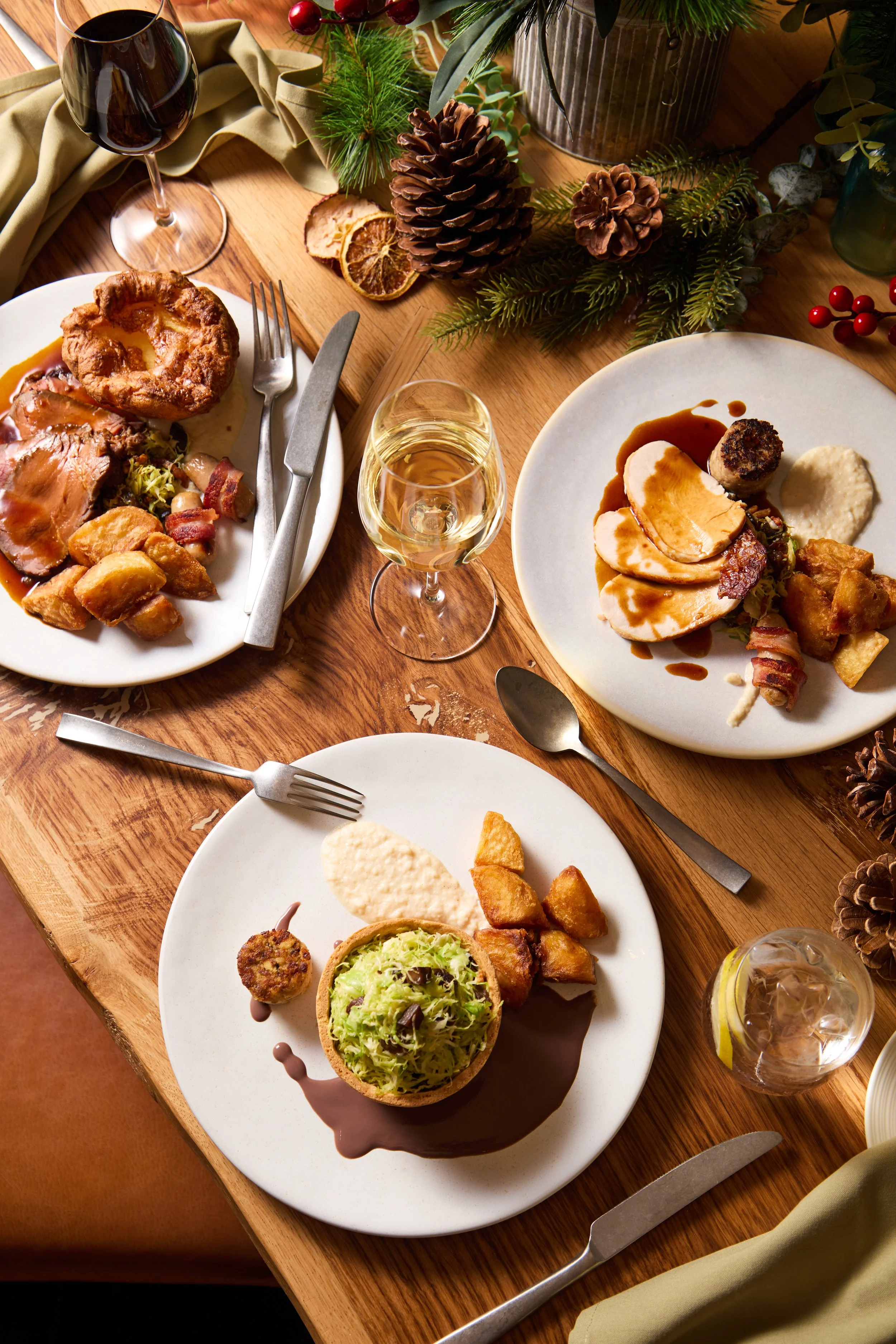 A festive holiday meal featuring plates of roasted turkey with gravy, stuffing, and roasted potatoes, alongside a salad with avocado, and bowls of food on a wooden table decorated with pinecones, berries, greenery, and glasses of red and white wine.