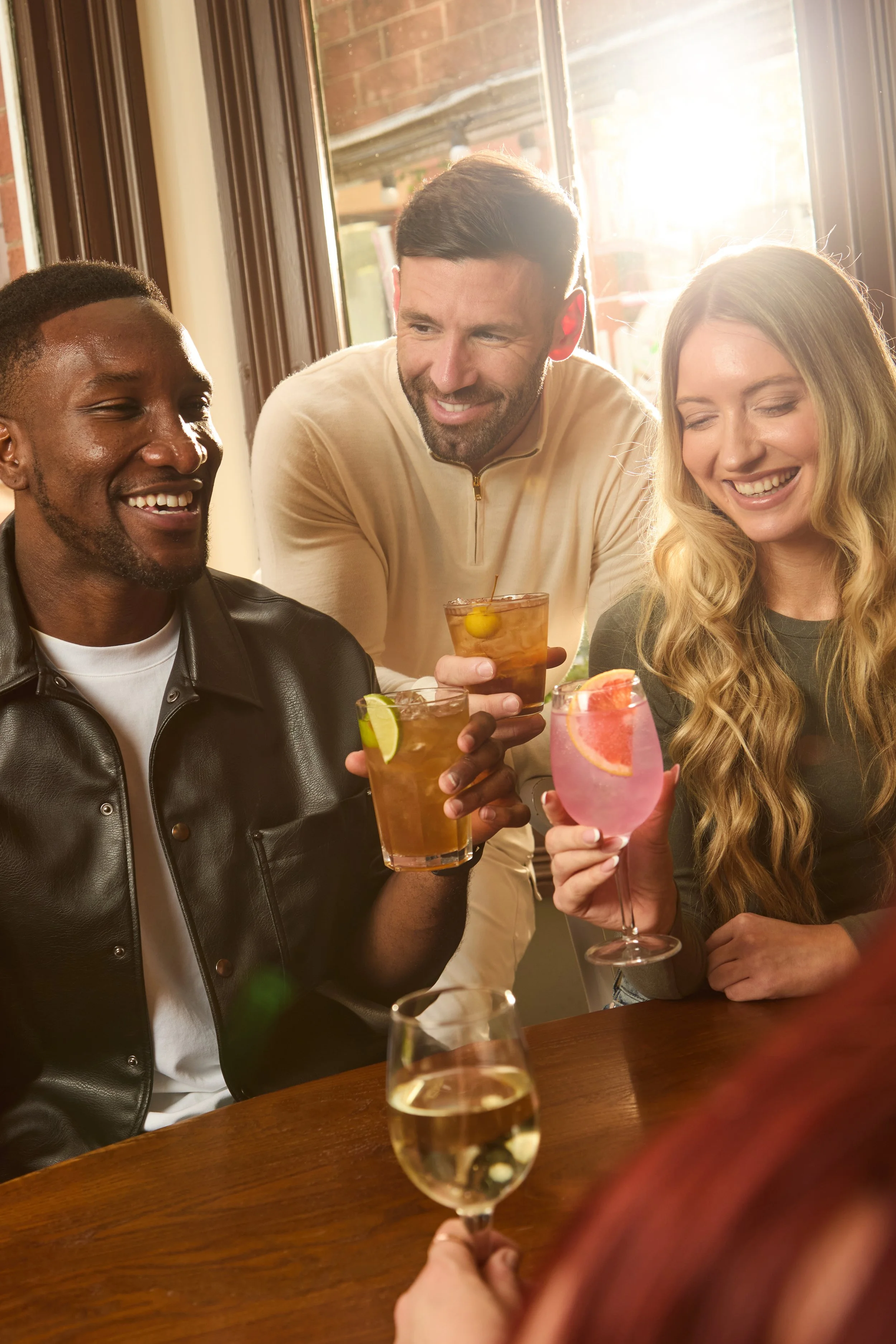 Three friends enjoying drinks together in a warmly lit restaurant or bar, smiling and toasting, with sunlight streaming through the window.