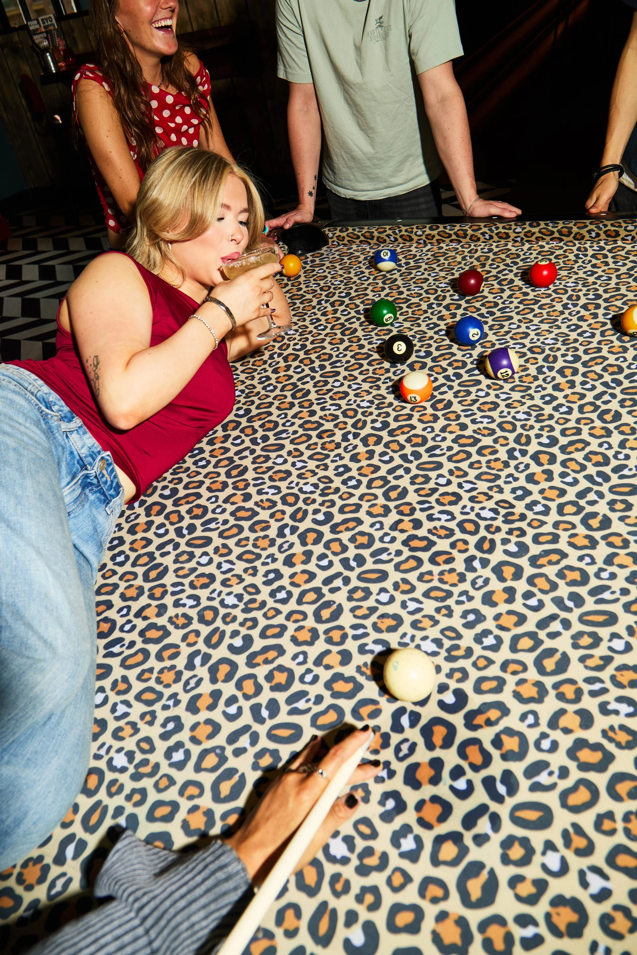 Four people playing pool on a leopard print table, with one woman in a red tank top drinking a cocktail, and others standing around.