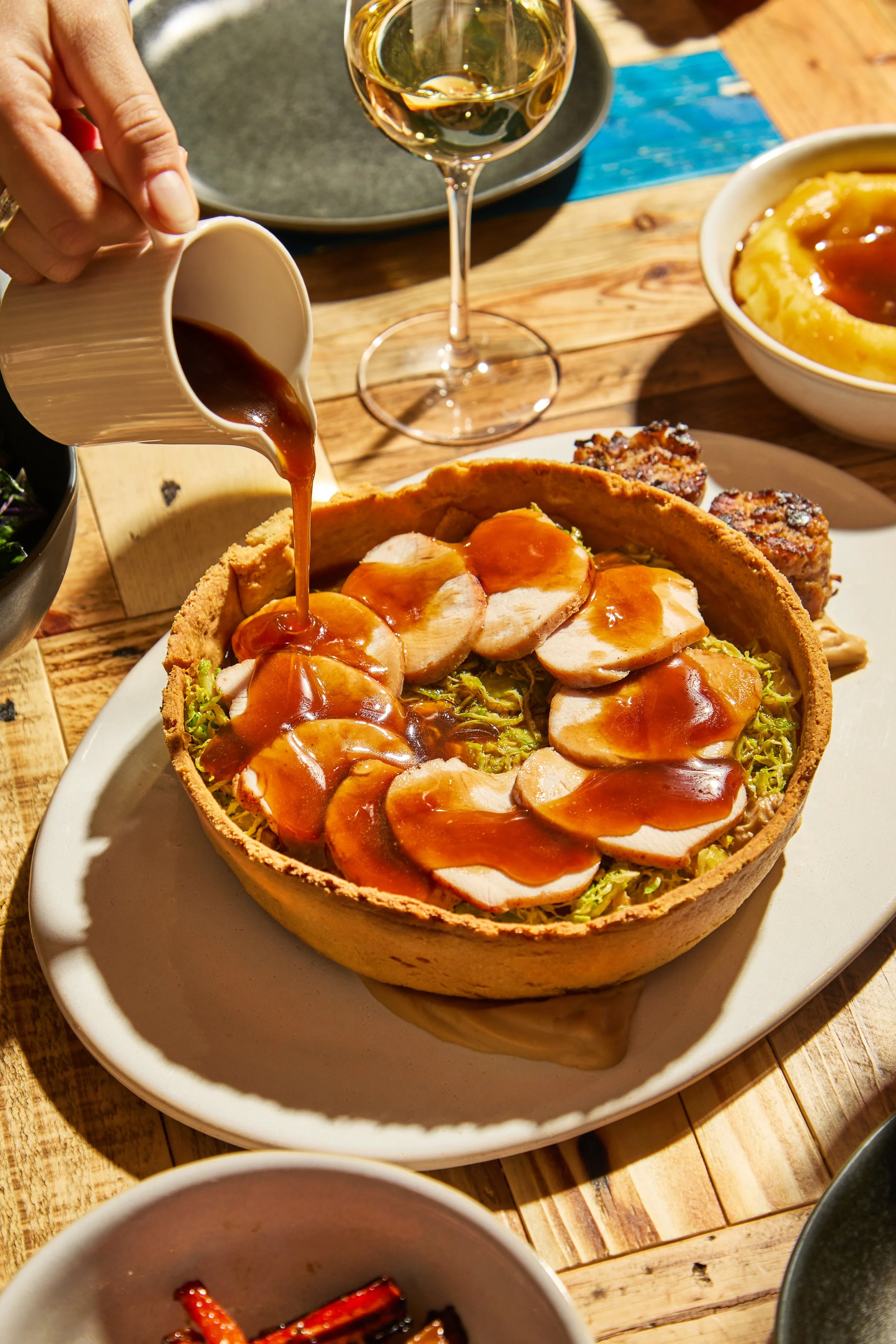 A pie with sliced turkey meat, shredded lettuce, and gravy being poured over it, on a white plate on a wooden table. There are side dishes and a glass of white wine in the background.