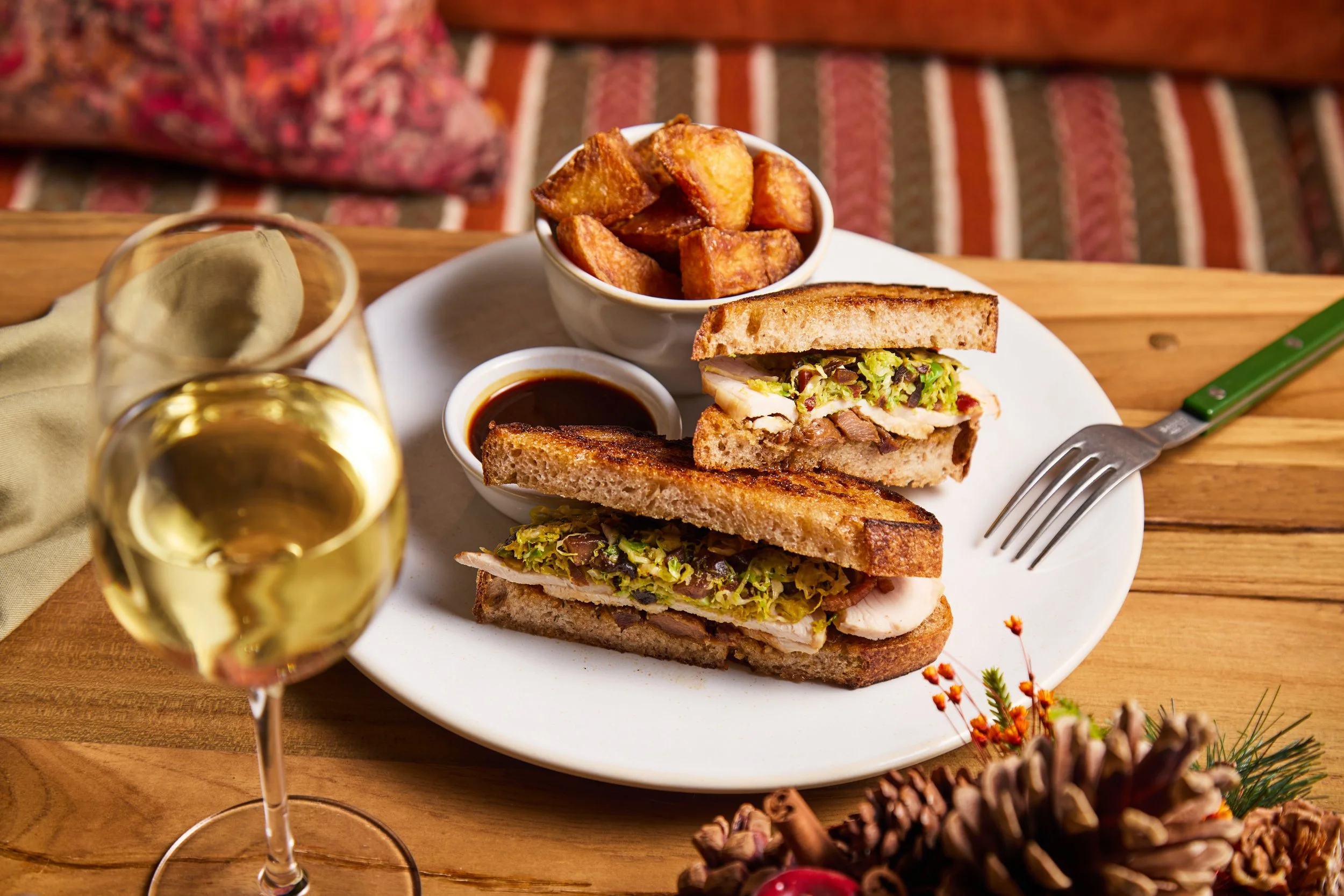 A plate with a sandwich, potato cubes, and dipping sauce, two glasses of white wine, and a fork on a wooden table.