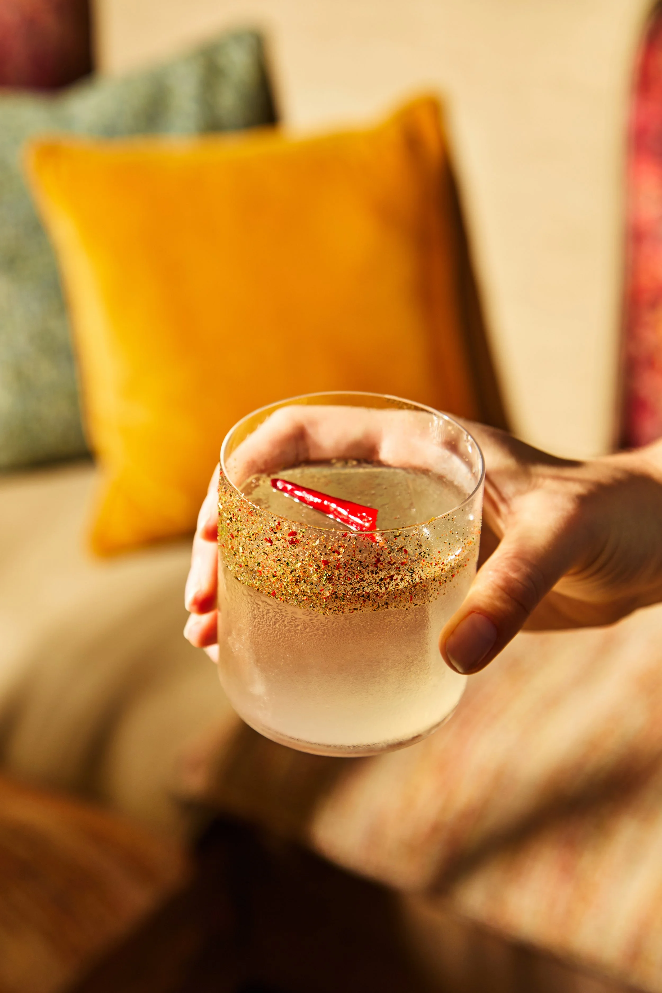 A hand holding a glass of a clear beverage with a red straw, decorated with a gold glittery festive ring around the rim.