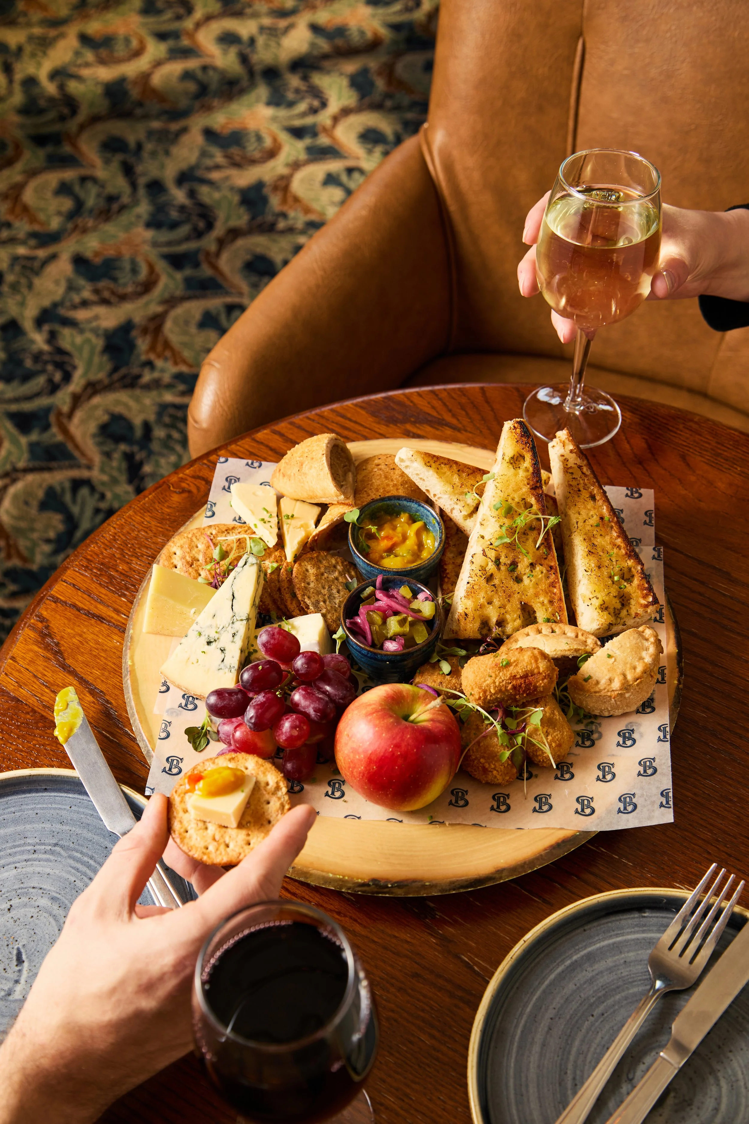A person holding a piece of toast with cheese and a boiled egg, surrounded by a cheese platter with grapes, apples, dips, and bread, with two glasses of red wine on a wooden table.