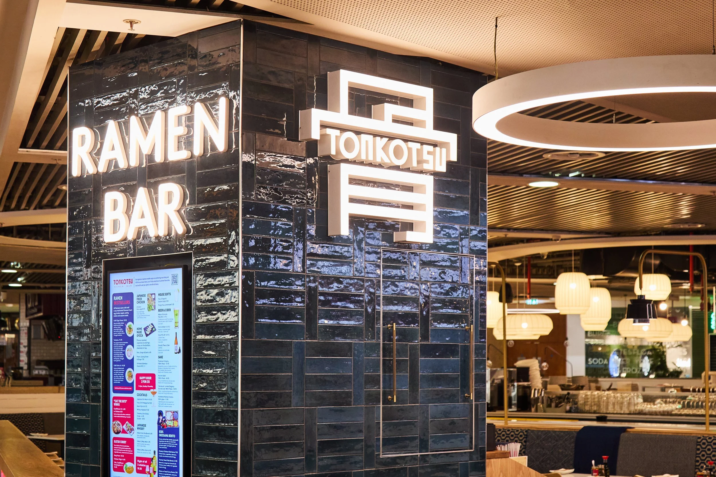 Interior of a restaurant with illuminated signs that read 'RAMEN BAR' and 'TOKOTSU'. The restaurant has dark tiled walls and modern lighting fixtures.