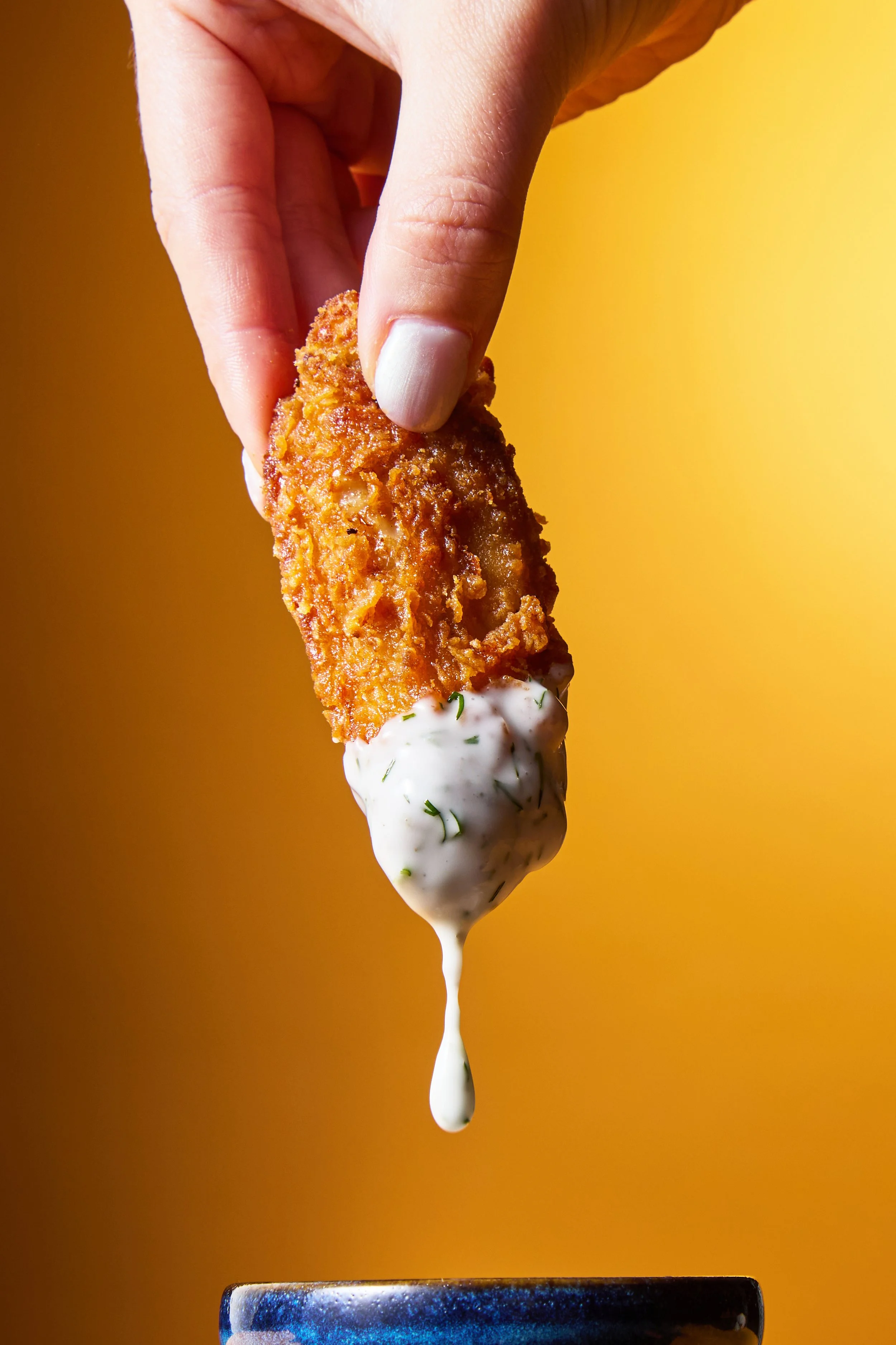 A hand holding a fried chicken tender dipped in creamy white sauce over a small blue bowl.