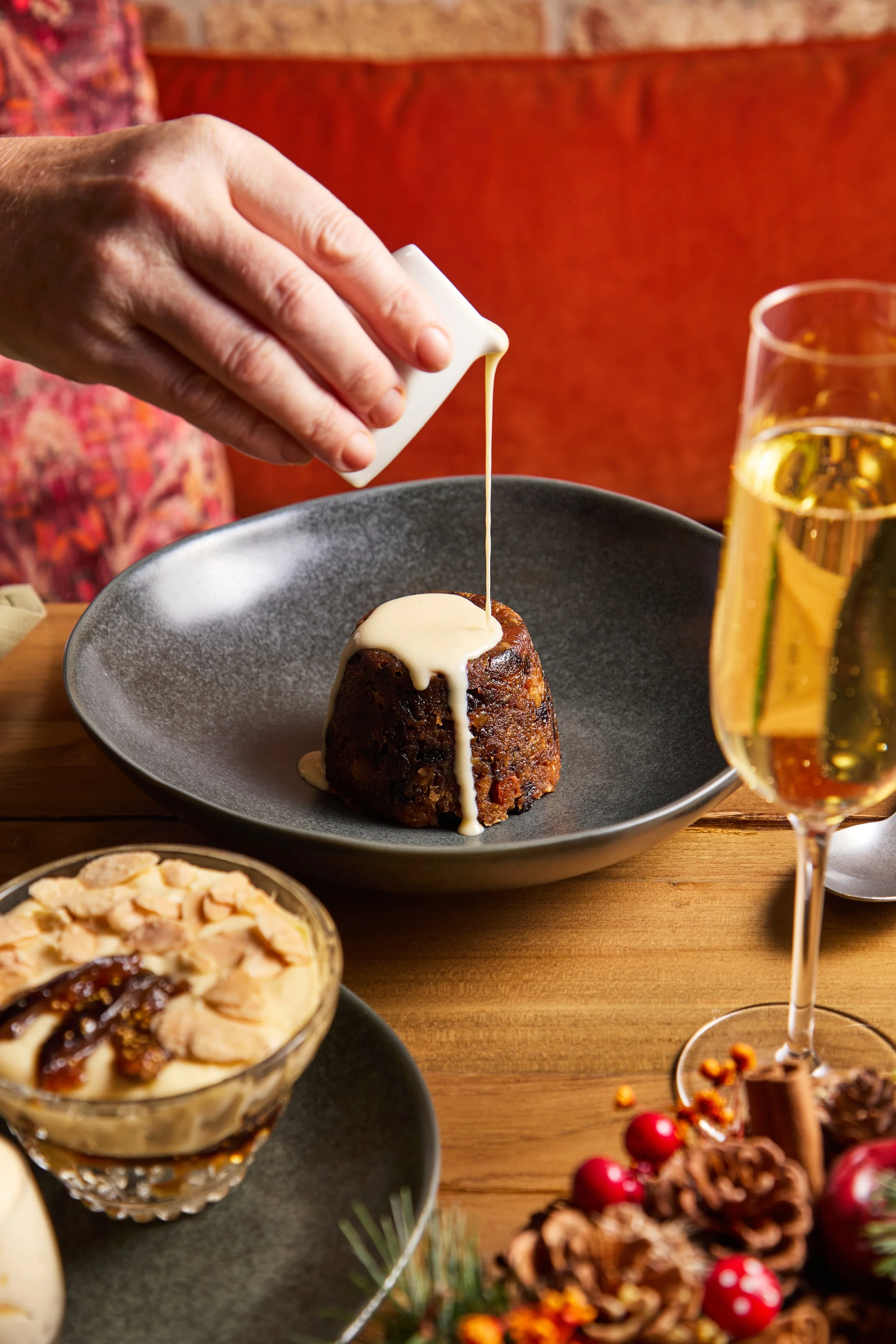 A dessert being topped with a white substance from a small pitcher, white syrup pouring over a chocolate cake in a black bowl, with a glass of white wine, a bowl of pudding, and a festive centerpiece with pinecones and red berries on a wooden table.