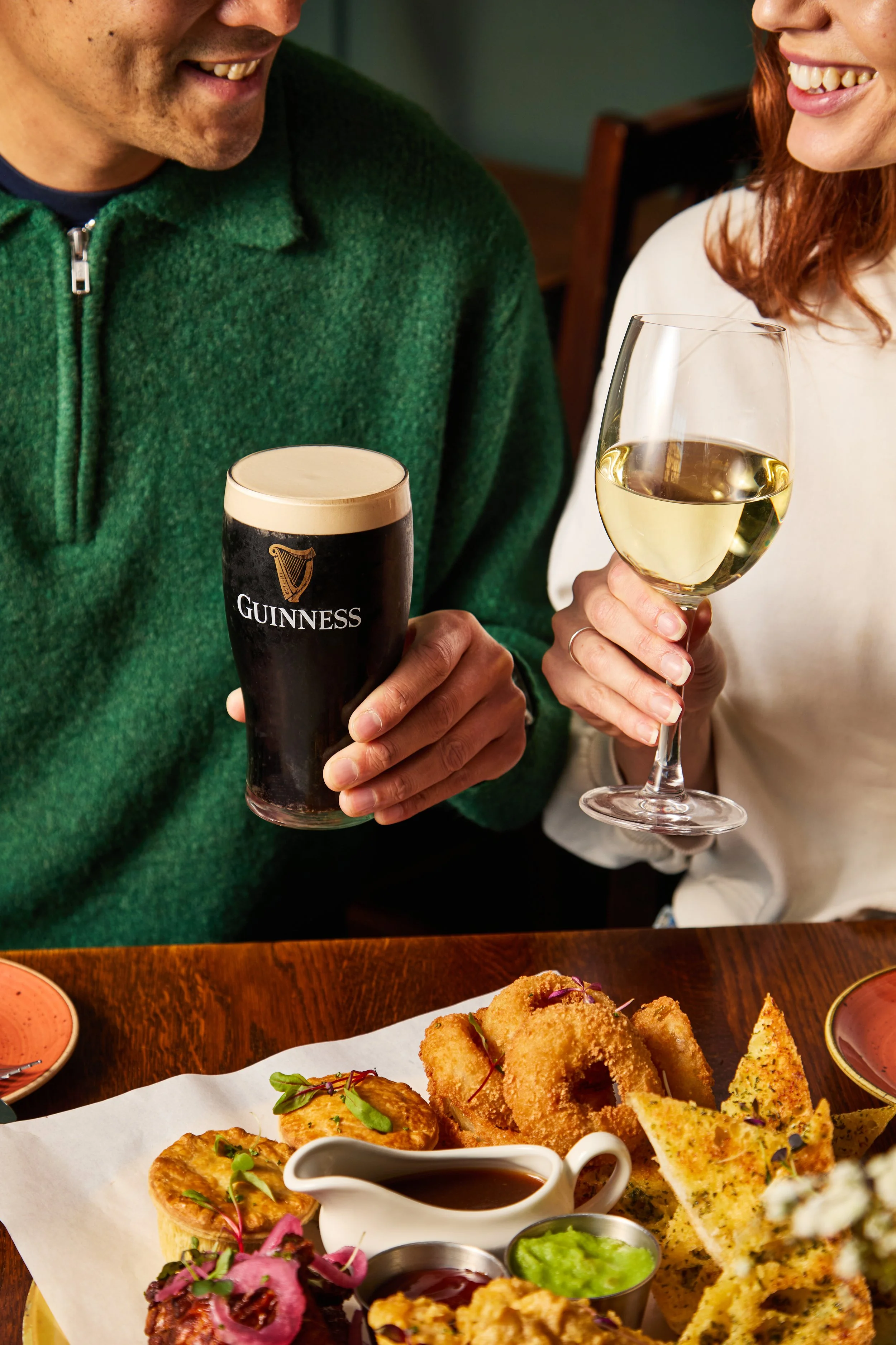 People smiling and clinking glasses of Guinness beer and white wine at a restaurant table with assorted appetizers.