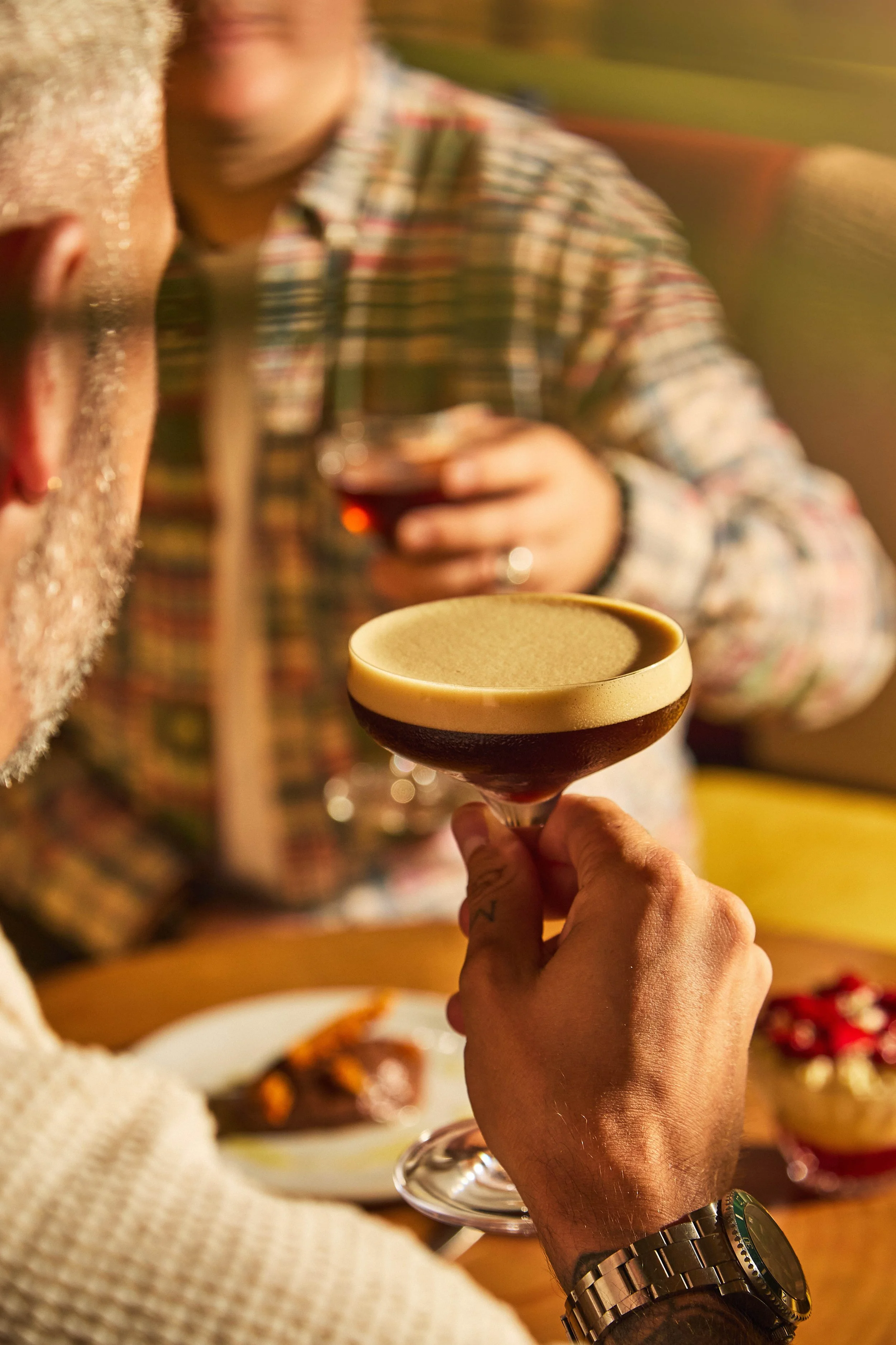 A person holding a cocktail glass with a dark drink inside. In the background, a person wearing a plaid shirt holds a glass of red wine. The setting appears to be a social gathering or dinner.