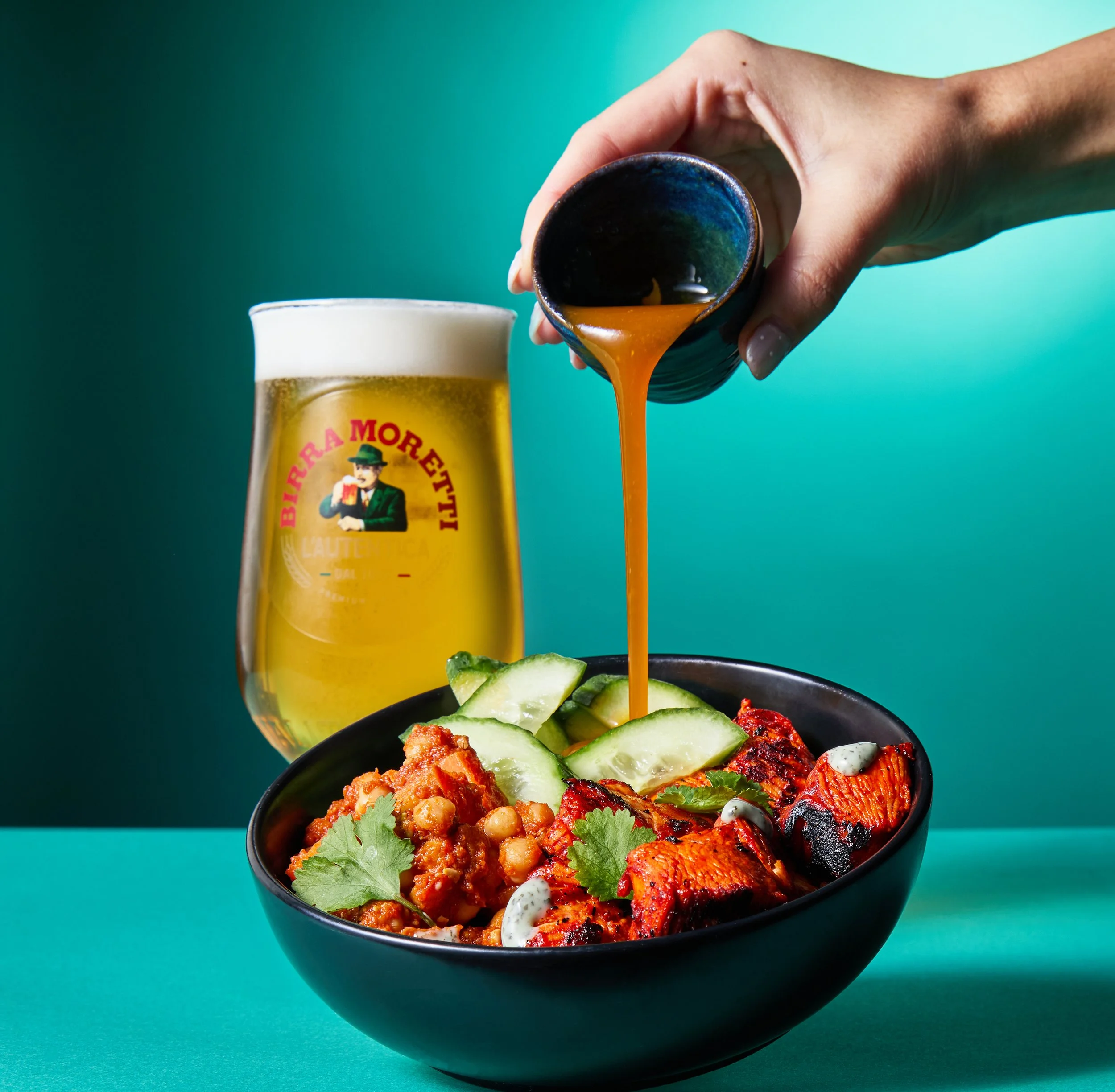 A bowl of Indian food with chicken tandoori, cucumber slices, chickpea curry, and cilantro, with a side of beer and a hand pouring sauce over the dish.
