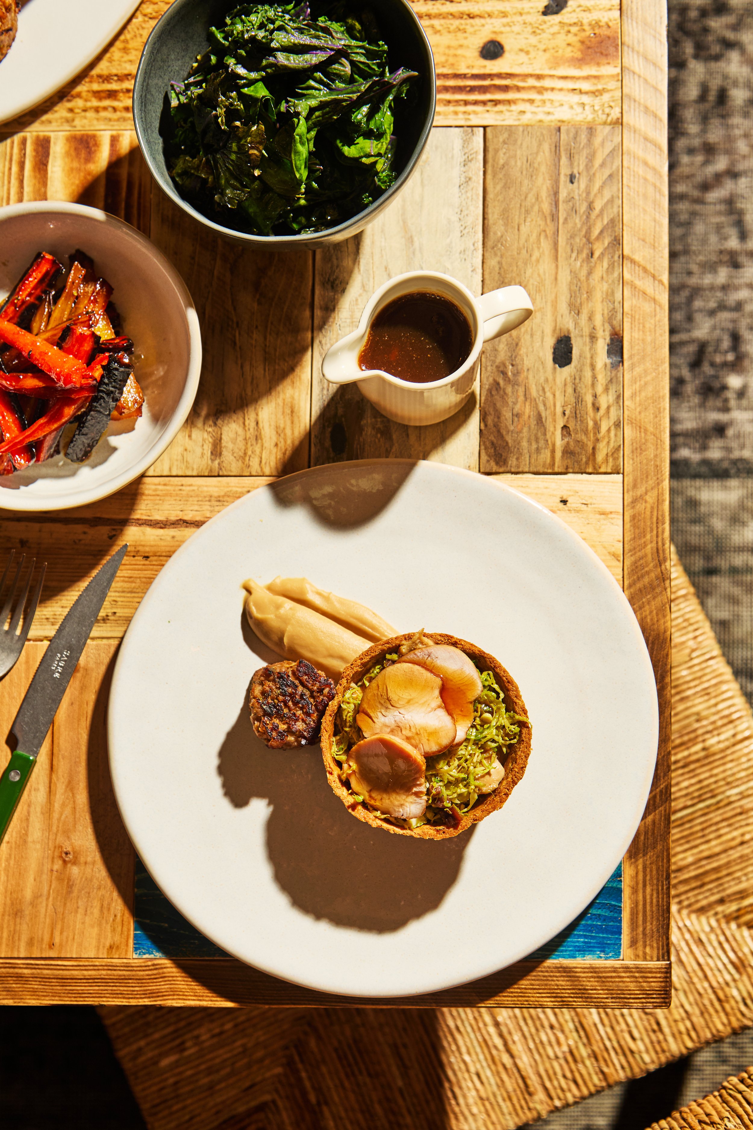 A plate with a meatloaf muffin, mashed potatoes, and a serving of shredded lettuce or cabbage salad, surrounded by side dishes, including a bowl of leafy greens, a dish of cooked vegetables, and a small pitcher of gravy or sauce, on a wooden table.