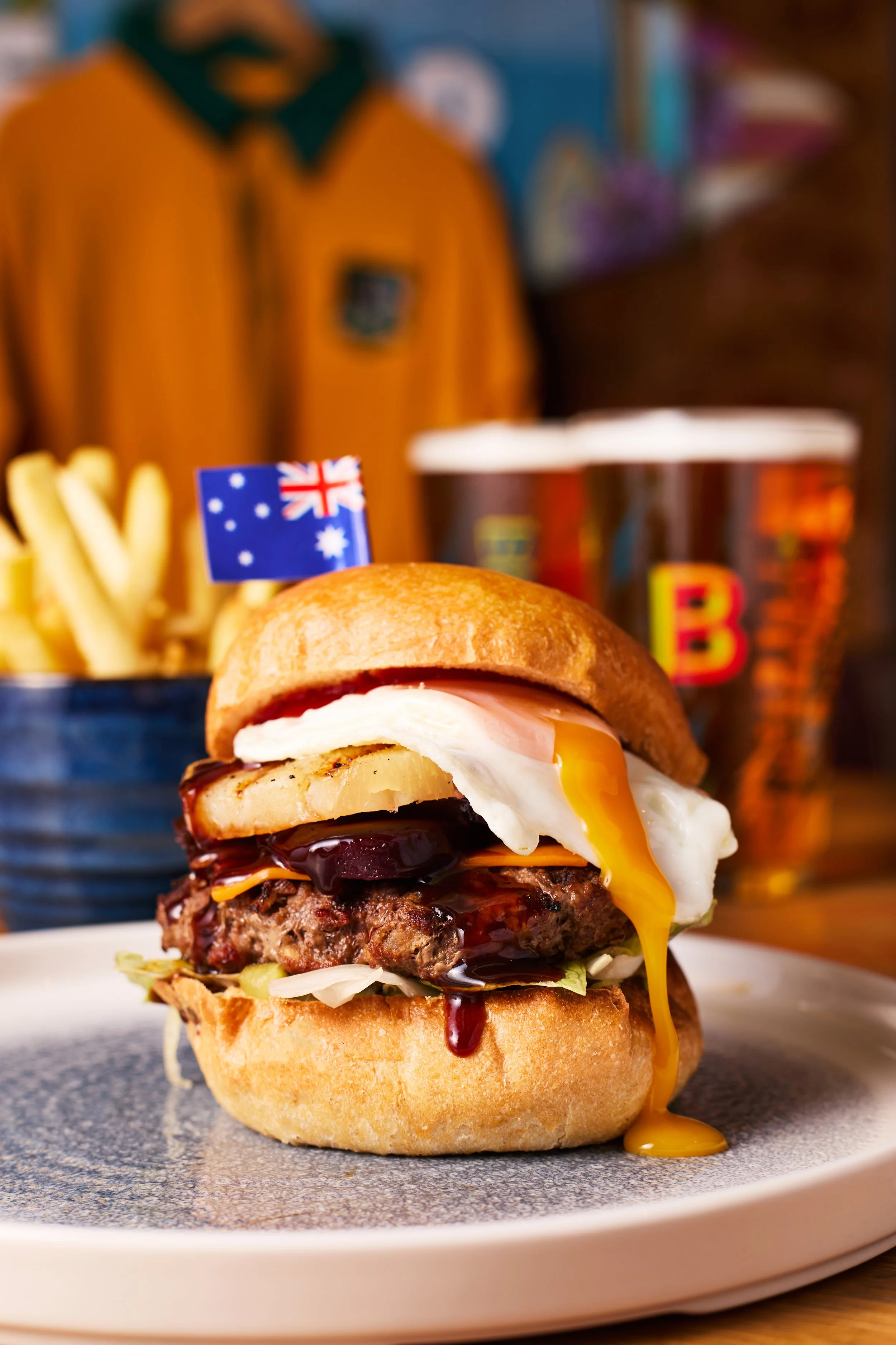 Close-up of a gourmet burger with an Australian flag toothpick, topped with a fried egg, pineapple, bacon, and cheese, served on a white plate with fries and drinks in the background.