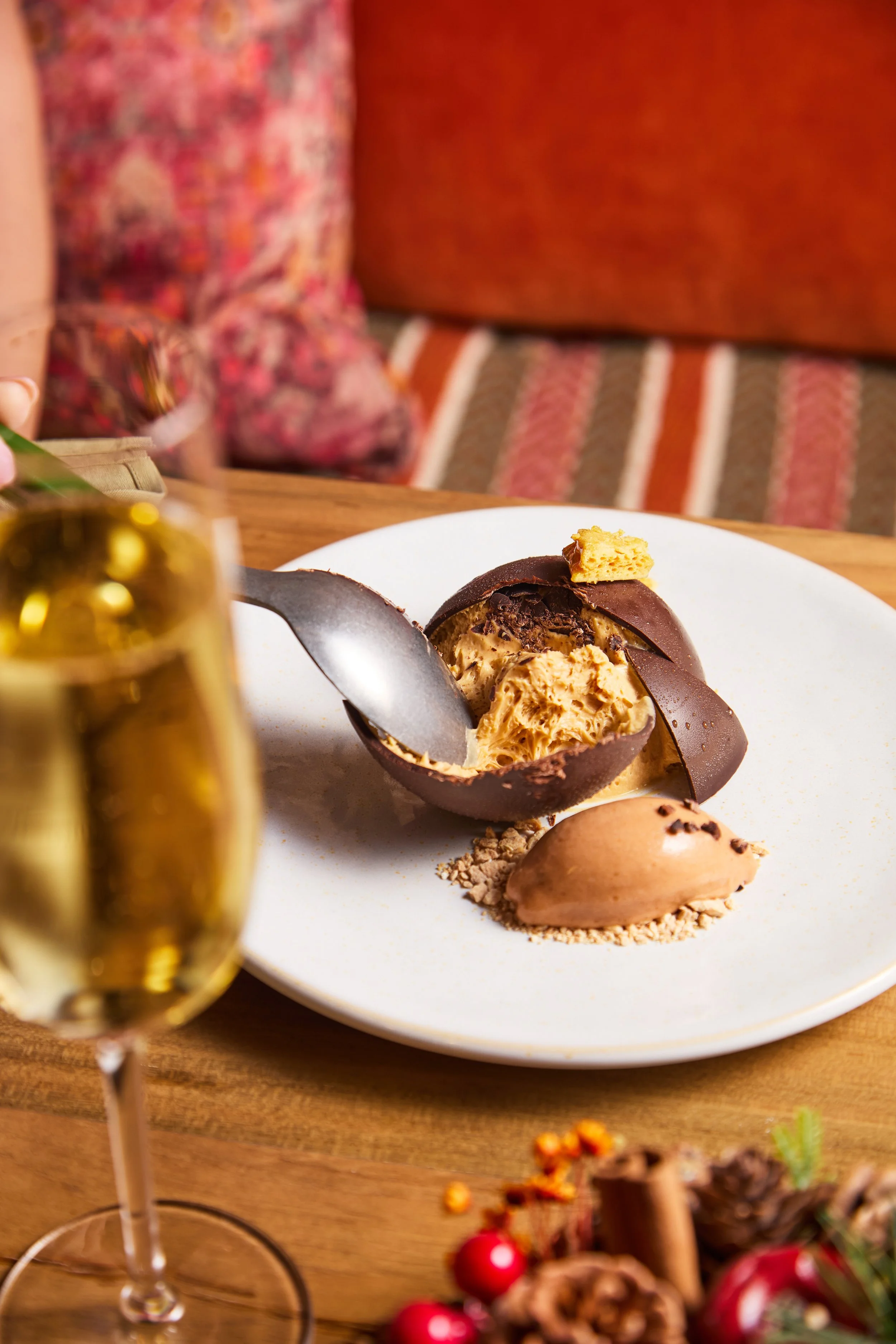 A white plate with a partially eaten chocolate and caramel dessert, a scoop of ice cream, and a spoon on a wooden table. In the foreground, a glass of champagne, and in the background, a cozy setting with patterned cushions.