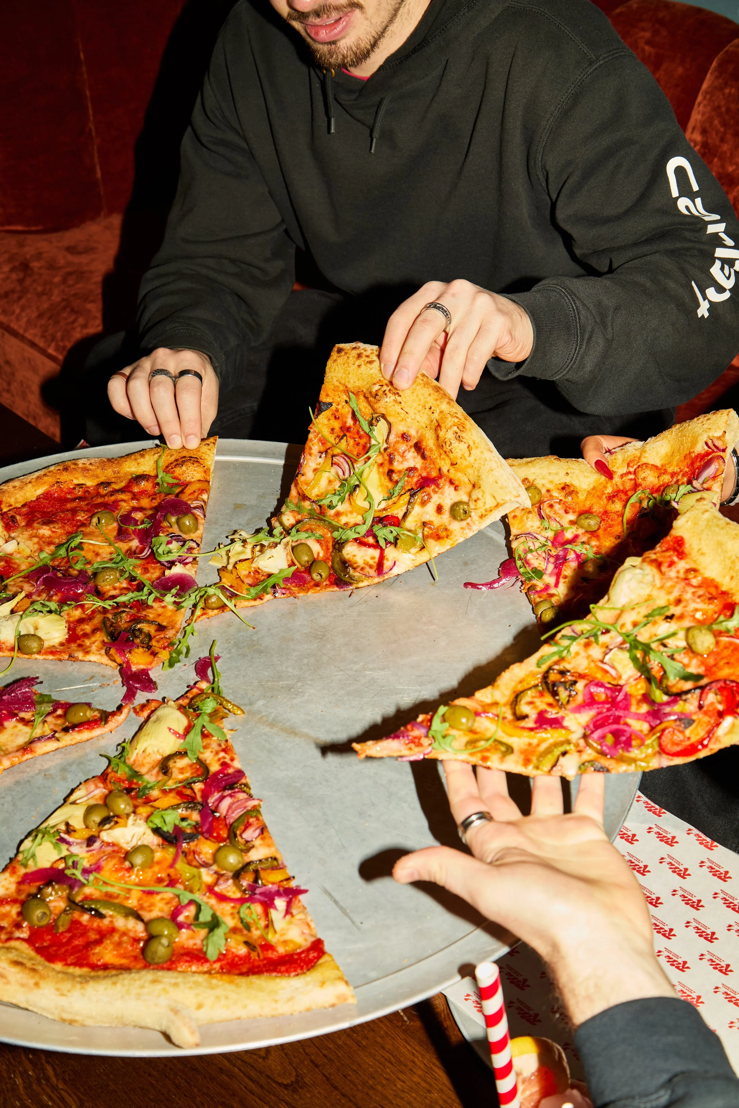 People reaching for slices of pizza on a large tray in a restaurant or pizza place. The pizza has various toppings including green olives, red onions, and greens.
