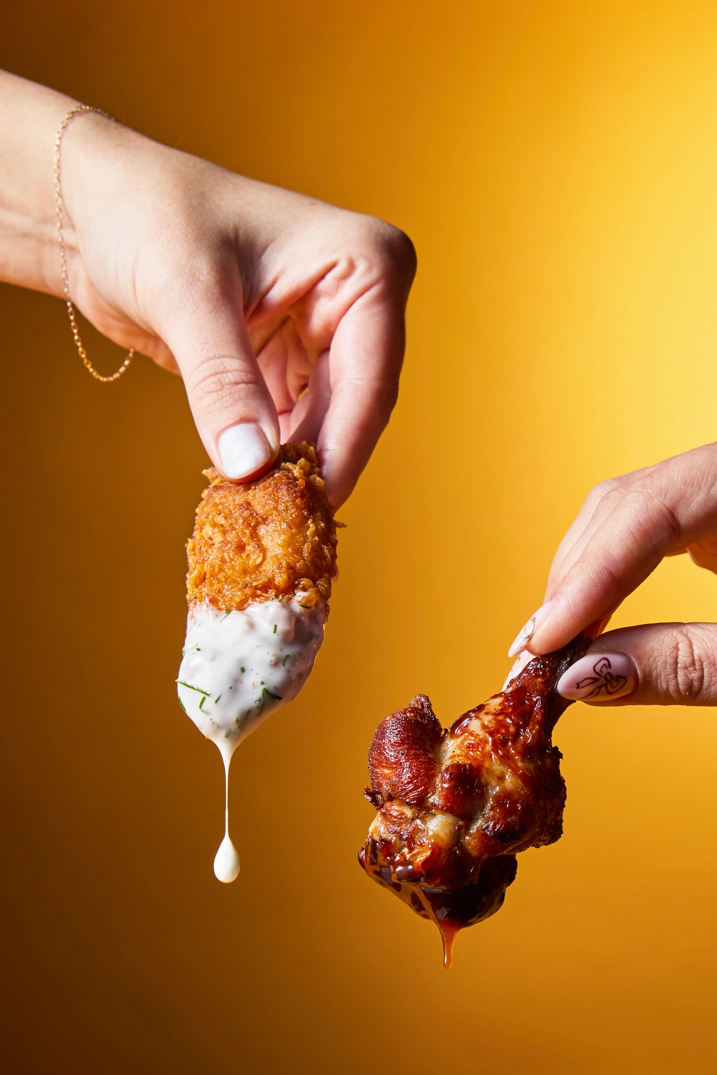Two hands holding different types of fried chicken drumsticks, one with white dipping sauce and the other with barbecue sauce, against a yellow background.