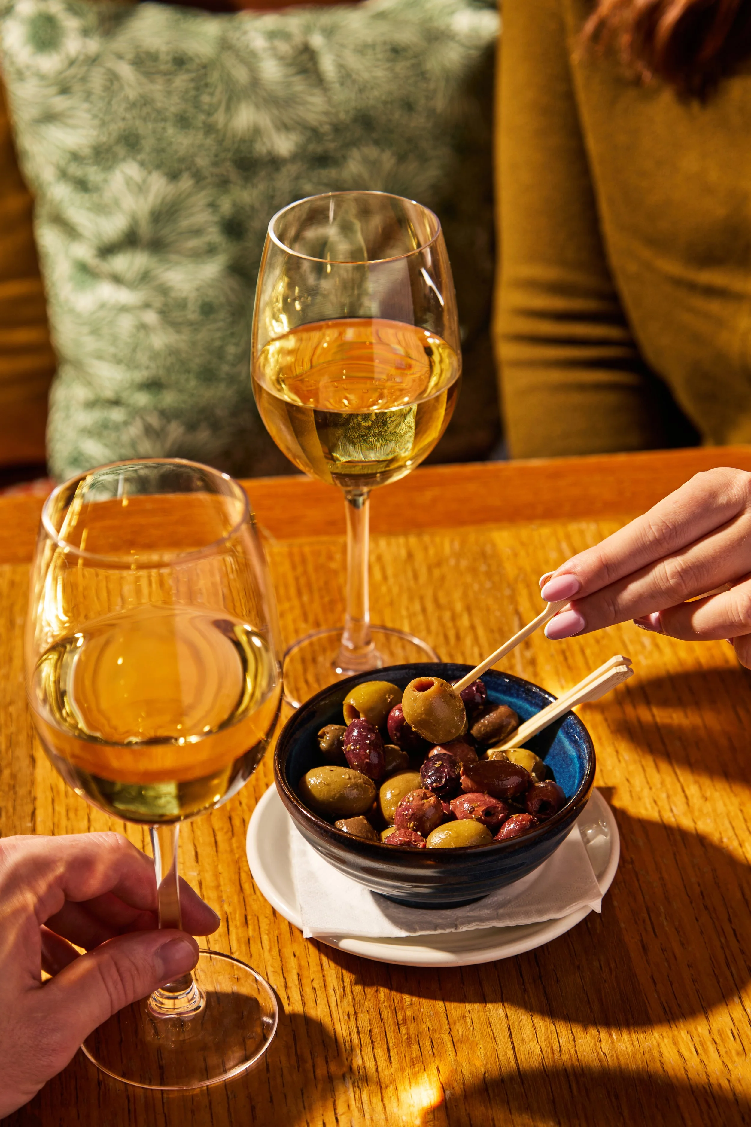 Two glasses of white wine, a bowl of assorted olives, and a person's hand picking an olive with a toothpick on a wooden table in a restaurant setting.