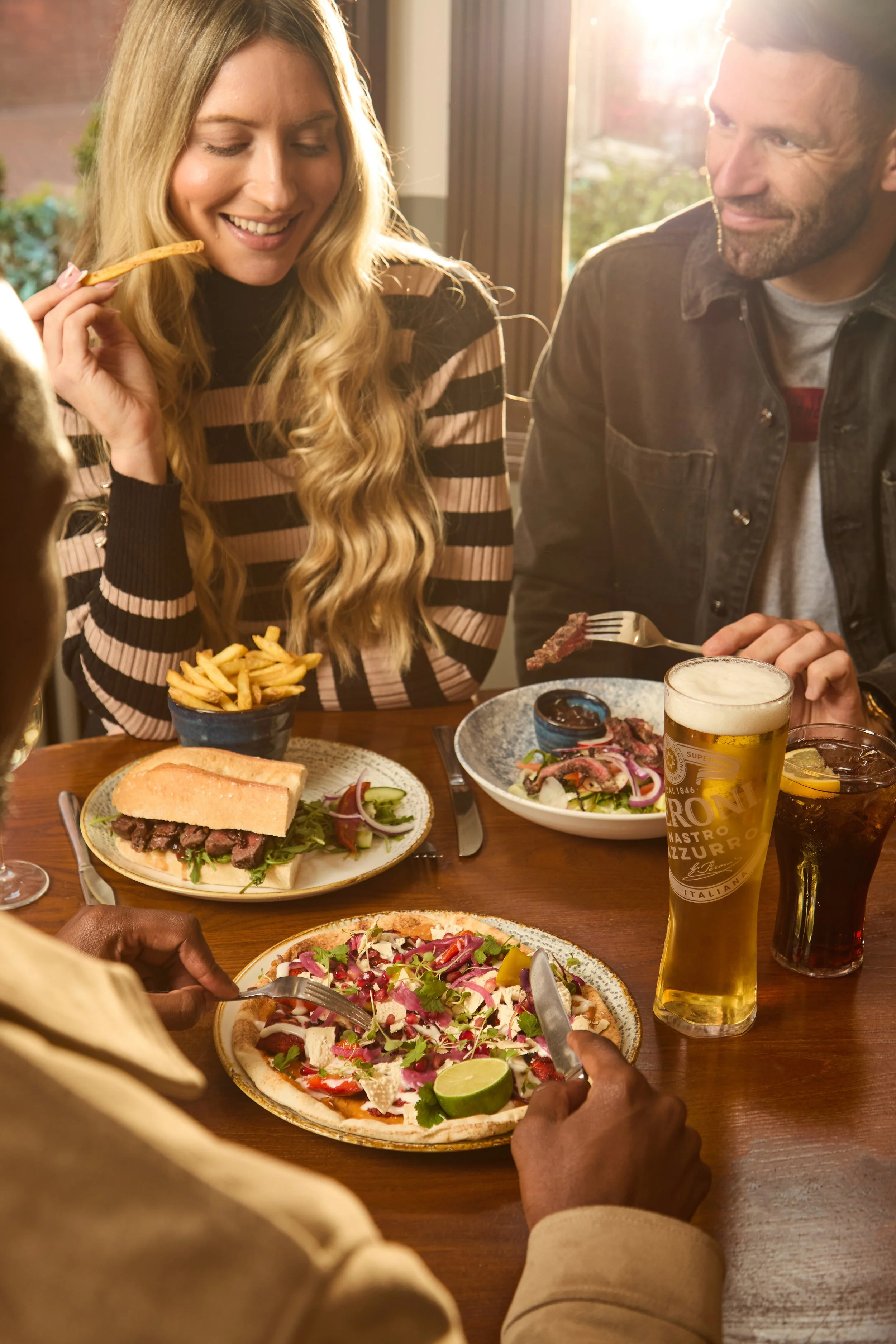 Group of people enjoying a meal at a restaurant, including pizza, salad, fries, and drinks on the table.