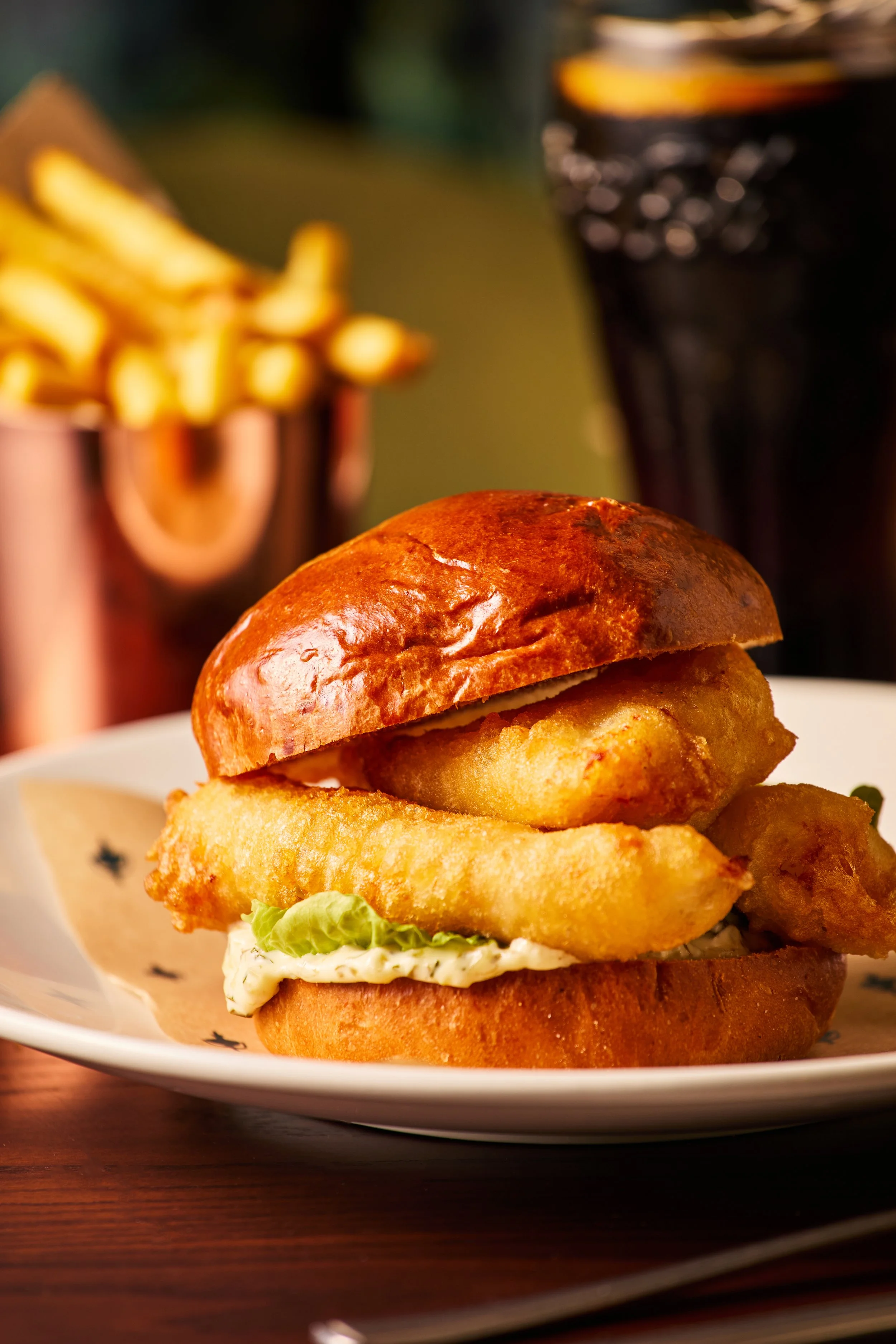 Close-up of a battered fish sandwich with a breaded fish fillet, shredded lettuce, tartar sauce, and a toasted bun, on a white plate with a wooden background.