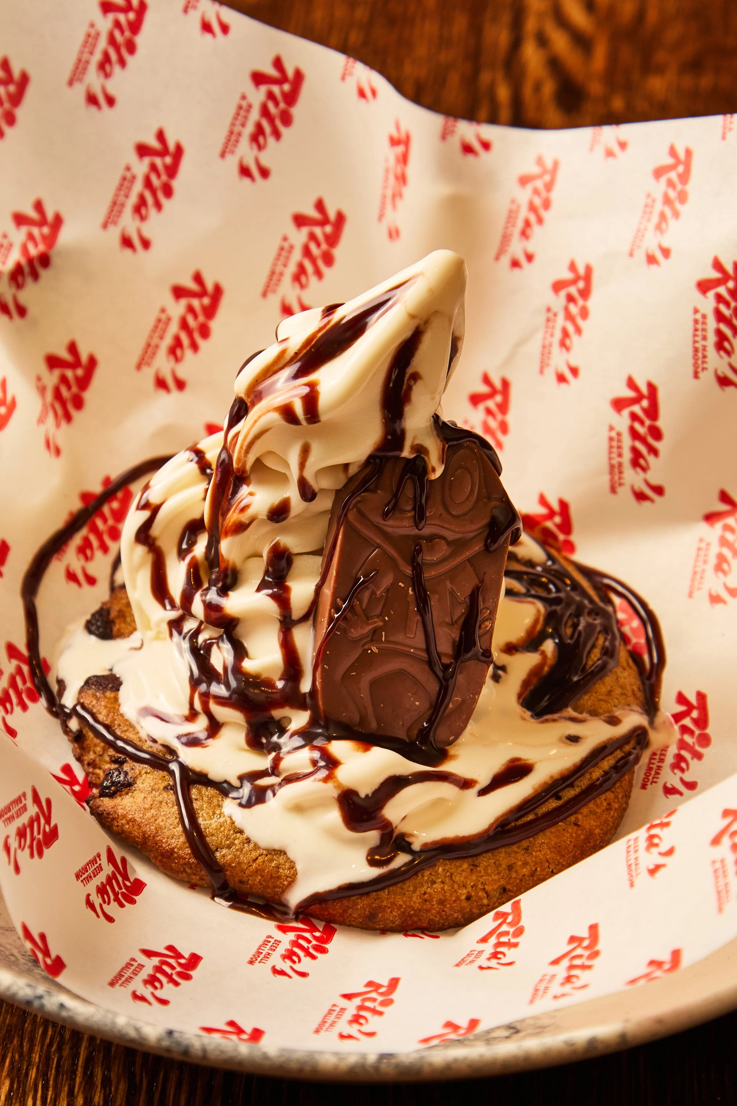 Chocolate chip cookie topped with vanilla ice cream, chocolate and caramel syrup, and a piece of chocolate, in a basket lined with branded paper.